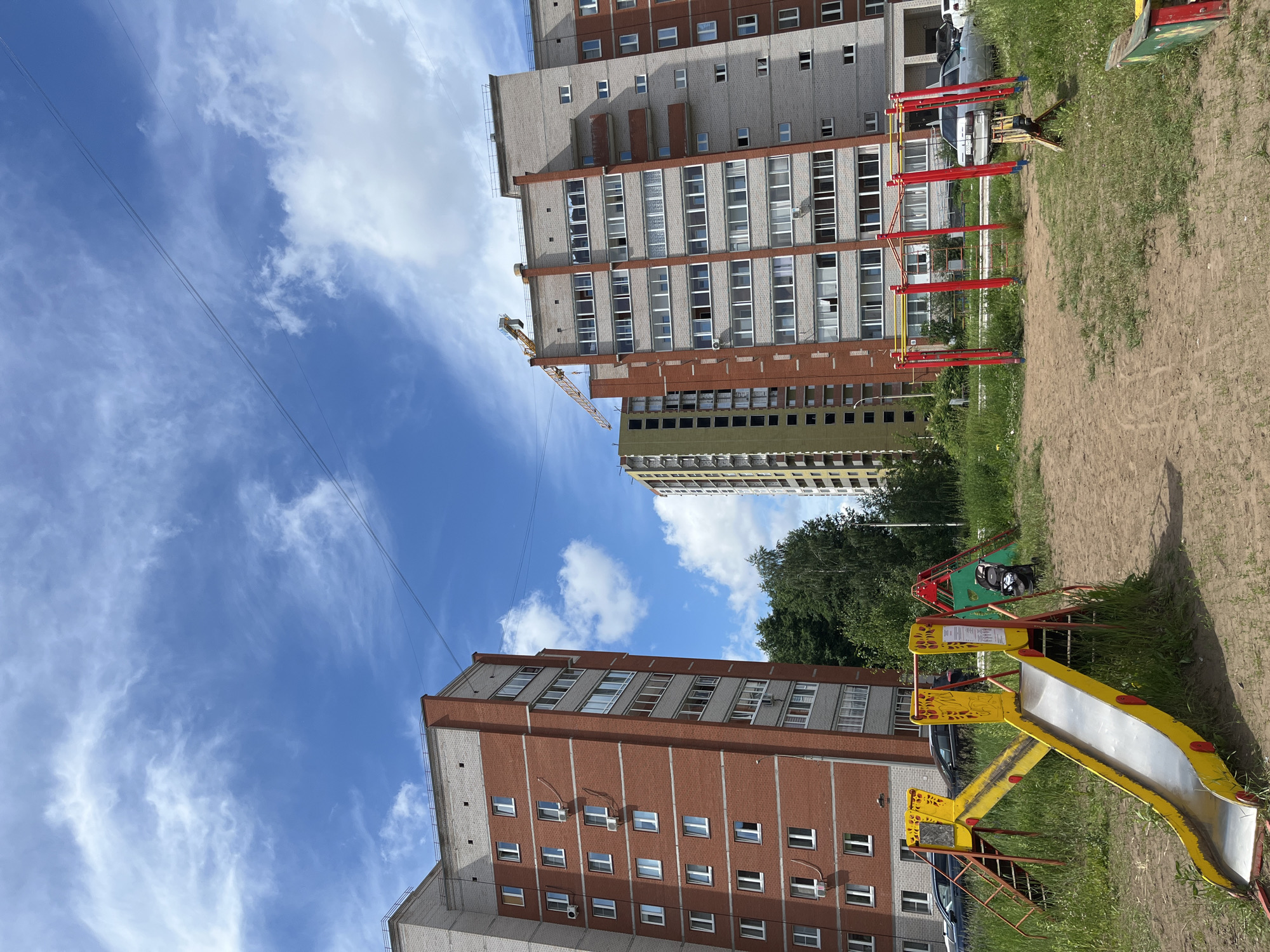 This is an outdoor, eye-level, full shot of a residential area in Chaikovsky, Russia, on a sunny day with a partly cloudy blue sky. In the foreground, a children's playground features a yellow metal slide with red supports and stairs, and a red metal swing set. The ground is a mixture of sand and grass. Behind the playground are two tall apartment buildings. The building on the left is constructed of red brick and light gray concrete. The building on the right is primarily light gray concrete with red brick accents. A third, unfinished building is visible in the background between the two completed buildings, with a construction crane extending from its top. Several cars are parked in the area between the playground and the buildings. Green trees are visible behind the playground and between the buildings. Power lines are strung across the sky.