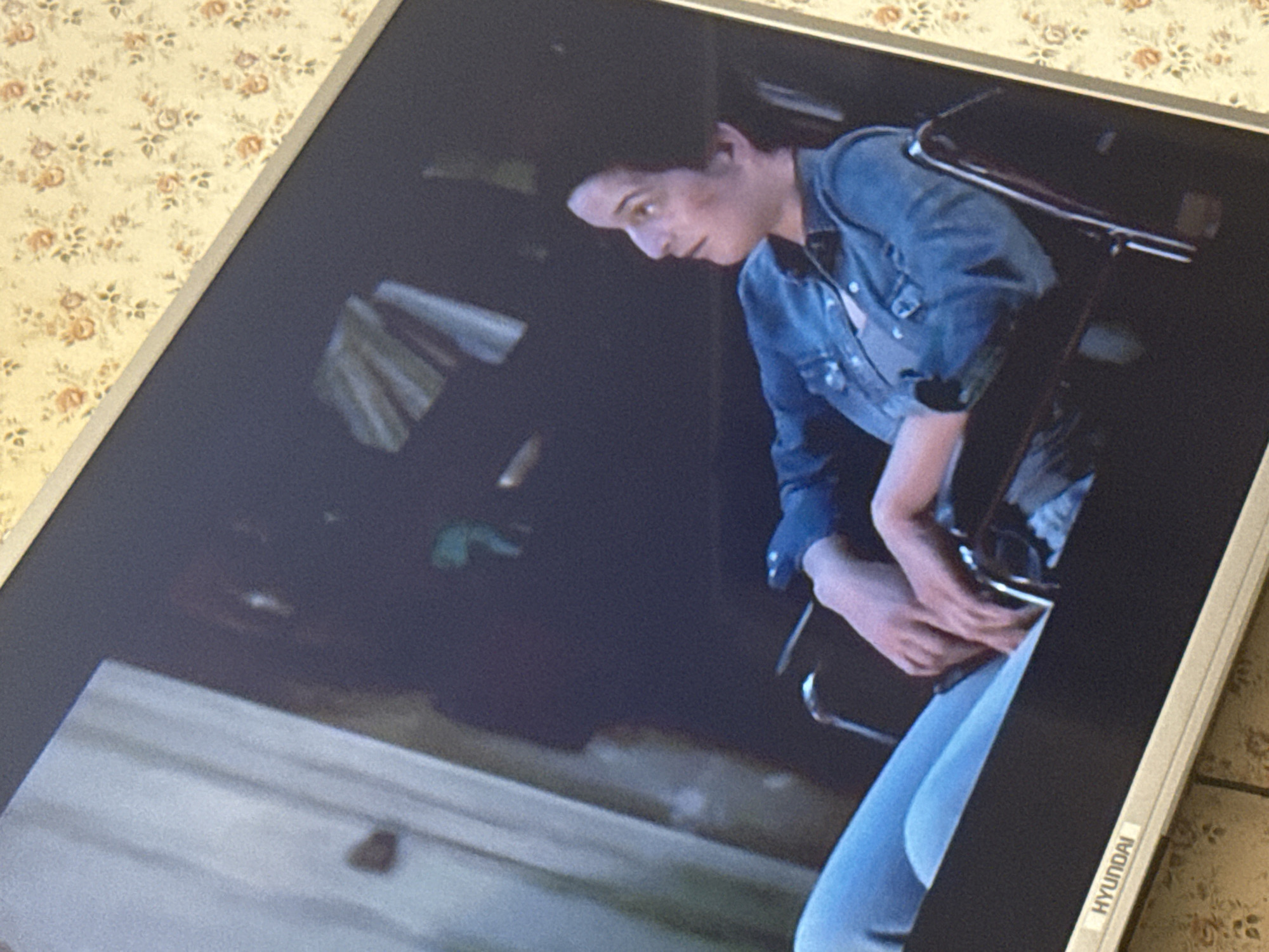 The image shows a Hyundai monitor displaying a scene with a woman in a denim jacket, seated, with a serious expression. The background is dark and indistinct, but suggests an indoor or enclosed space. The screen itself is resting on a surface with a floral pattern. The location is not discernible from this image.