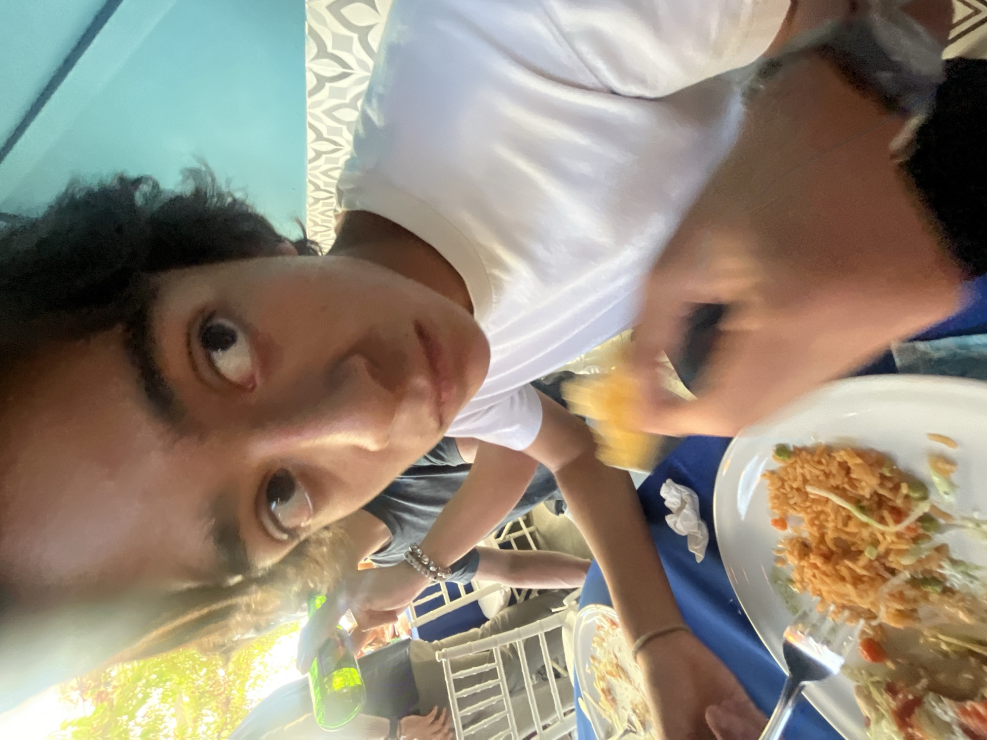 The image appears to be a selfie taken at an outdoor dining establishment, possibly in the city of Puebla, Mexico, based on the architectural patterns. The central figure, a person with dark hair, is wearing a white t-shirt. Other people are visible at the table and in the background. The dining area includes white plastic chairs and tables covered with blue tablecloths. Plates of food, including rice and other unidentified items, are on the tables. A bottle of what appears to be alcohol is also present. The photo is taken at an angle, with the subject looking towards the camera. Sunlight is visible in the background.