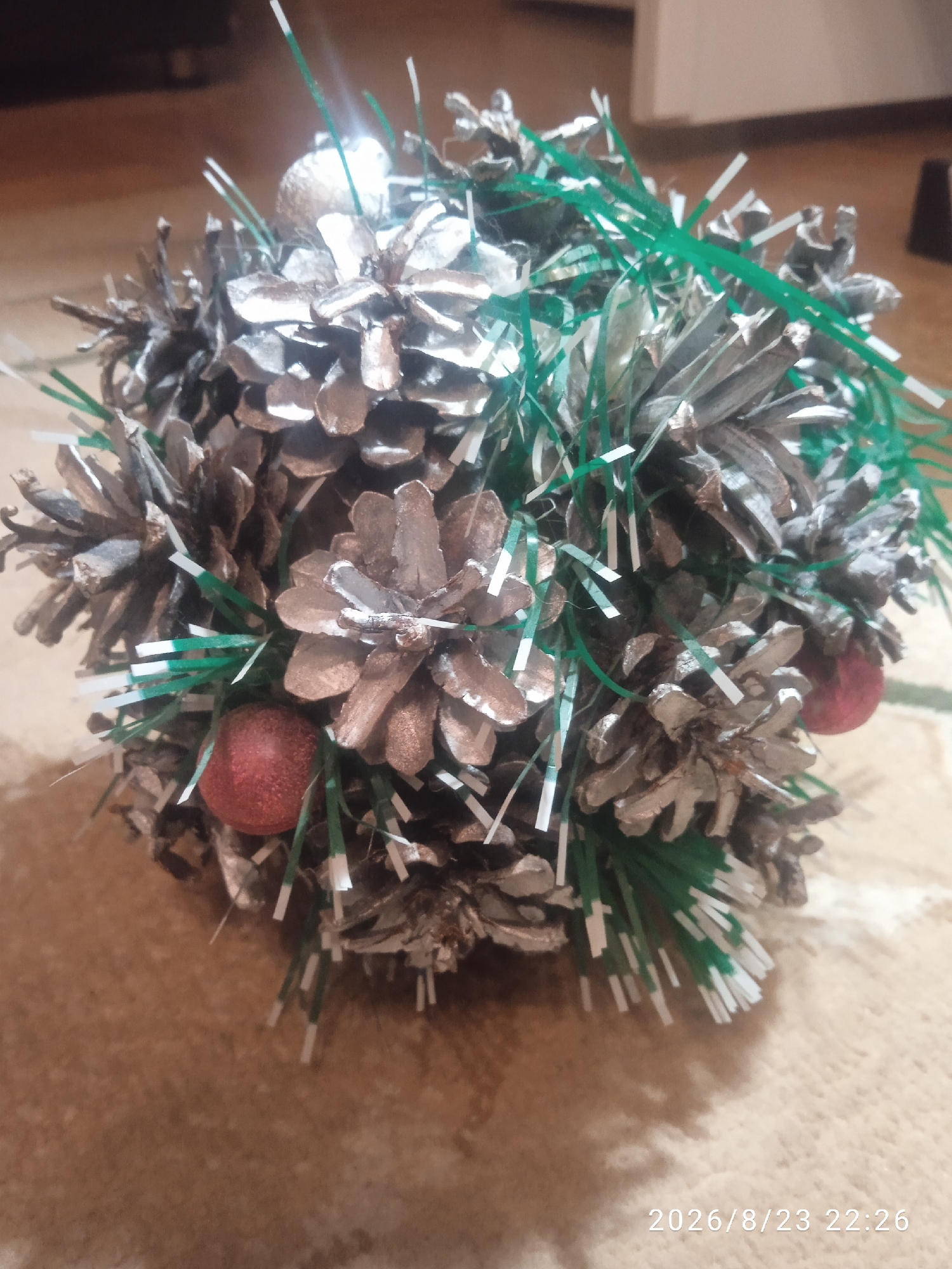 The image displays a close-up, slightly angled view of a decorative object made of pine cones, tinsel, and two small red spheres. The pine cones appear to have been painted or sprayed with a silver or metallic color, and they are interspersed with green and white tinsel. The object is spherical in shape and rests on a textured surface that resembles a woven mat or fabric. In the bottom right corner of the image, a timestamp is visible: "2026/8/23 22:26". The background is out of focus, showing what appears to be a wooden surface and a lighter colored wall or surface. There are no people or discernible actions in the image. The location within the city of Lida, Belarus, is not provided in the image itself.