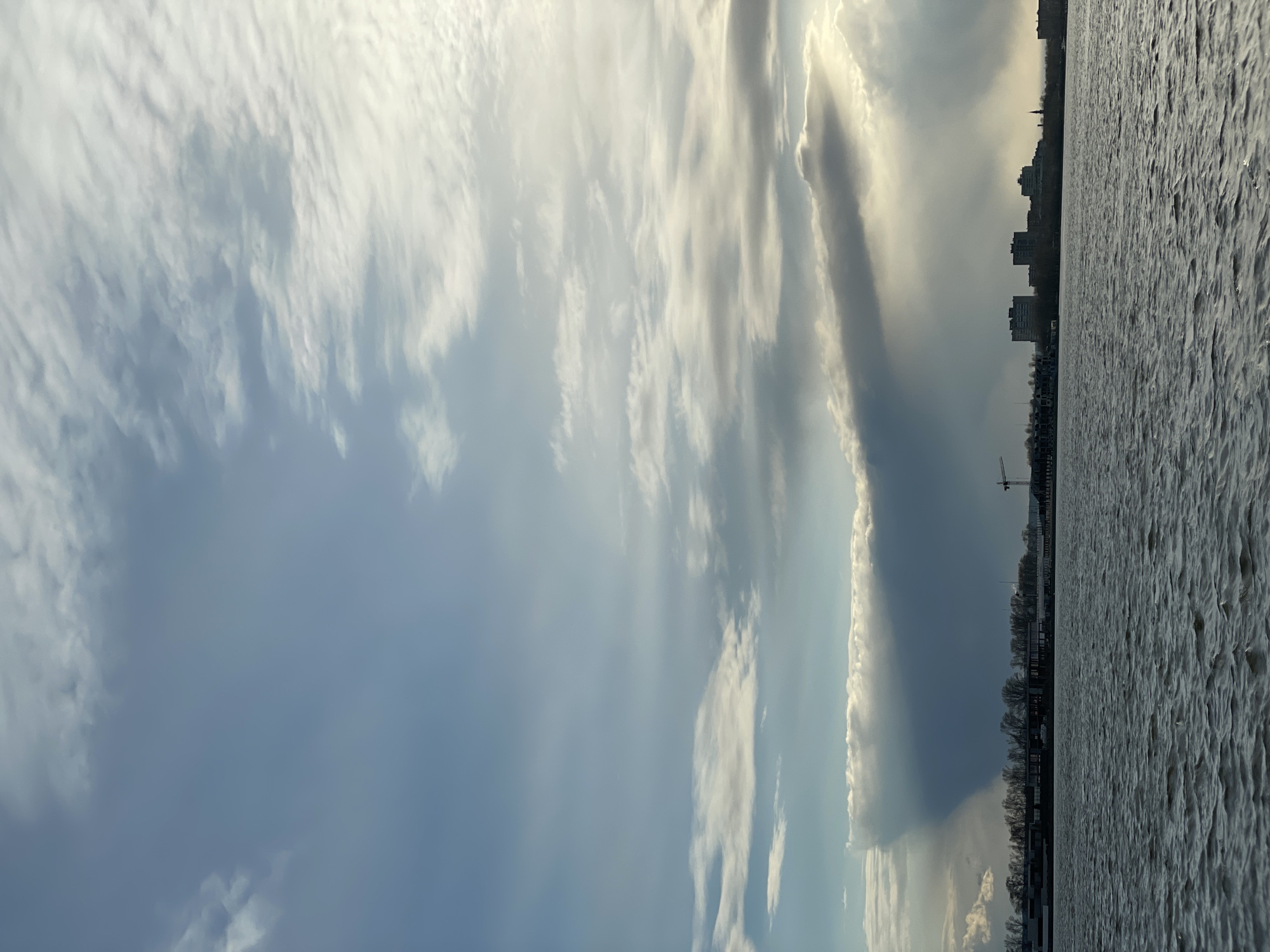 This wide landscape photograph captures a scenic view of a large body of water adjacent to a city skyline, all under a dramatic, expansive sky. The dominant feature is the sky, which fills the upper portion with a diverse array of clouds. Bright, wispy clouds are strongly illuminated by sunlight, creating prominent crepuscular rays that pierce through darker, heavier cloud formations. The sky displays a gradient of colors, ranging from brilliant white and soft golden where the sun breaks through, to a deeper blue-grey in areas of denser cloud cover.
Along the horizon, a distant city skyline is visible, featuring a series of multi-story buildings and a narrow strip of bare trees, all silhouetted against the bright sky. A tall construction crane is discernible near the water's edge on the far right of the urban strip.
The lower portion of the image is occupied by a vast expanse of water, likely a river or lake, characteristic of Kazan, Russia. The water's surface is textured with countless small ripples or waves, appearing a uniform silvery-grey, suggesting either movement or possibly very slight ice formation.
The lighting indicates either a late afternoon or early morning scene, with the sun low on the horizon, casting a dynamic interplay of light and shadow across the clouds. The absence of foliage on trees points to a winter or early spring setting. There are no people or discernible text in the image.