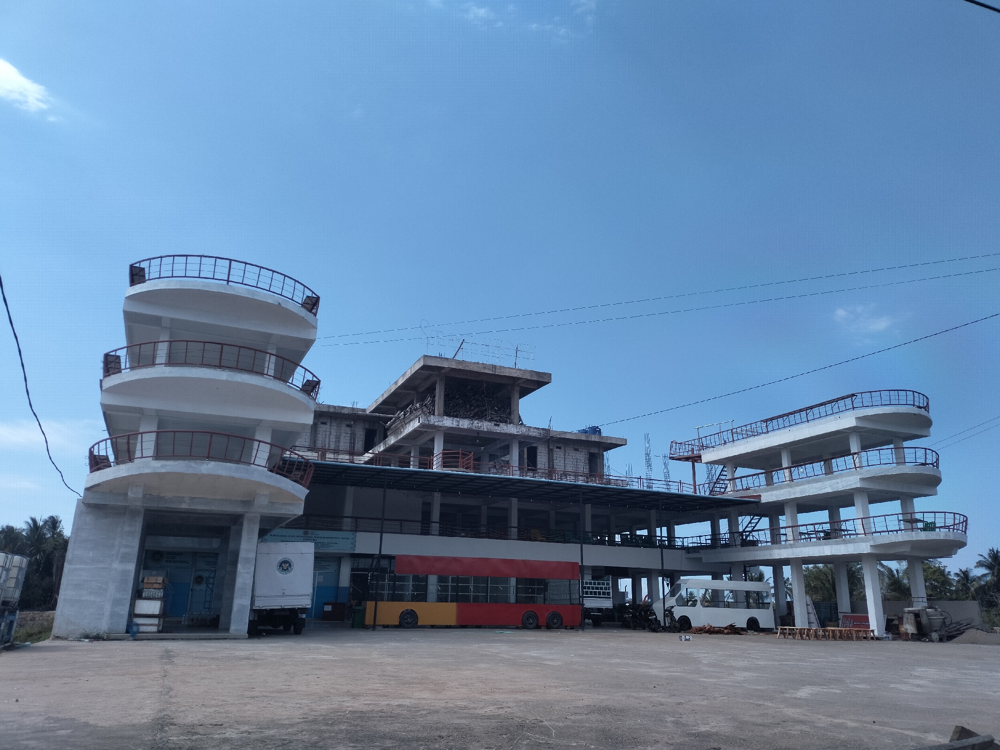The image depicts a large, partially constructed building in Teupinpukat, Indonesia. The architectural style is a mix of modern and functional, with several tower-like structures featuring multiple levels and rounded balconies. The central section of the building is still under construction, with exposed concrete and rebar visible.
Various vehicles are parked in front of the building, including a box truck, a bus, and a white van. The foreground is dominated by a large, open paved area. The sky is clear and blue. The surrounding environment includes trees. There is no sign of people present.