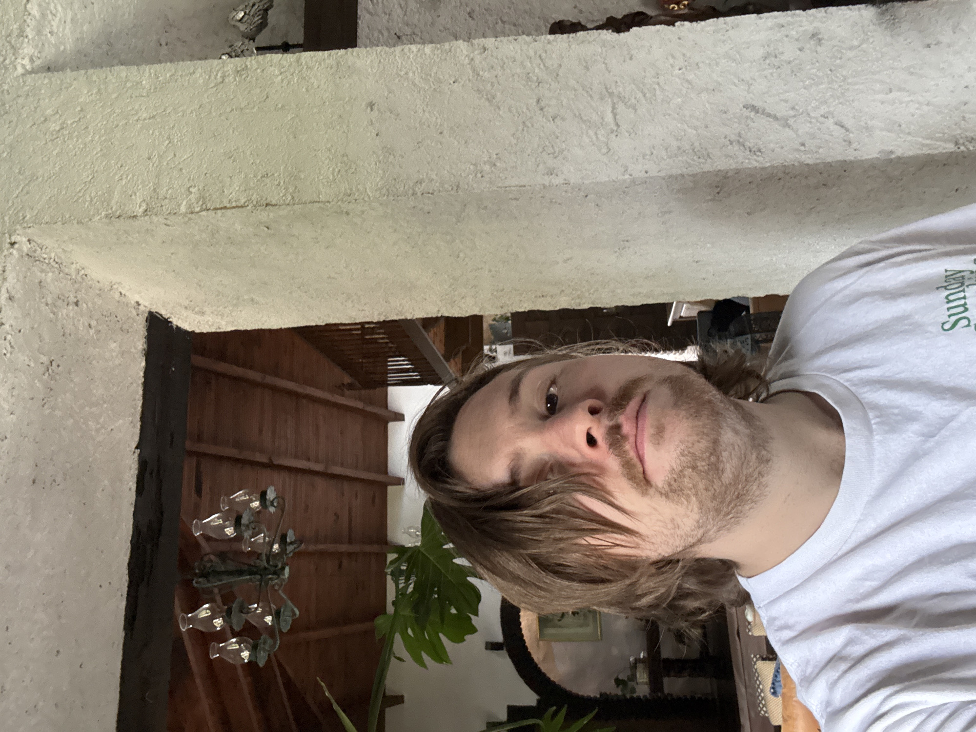 The image is a selfie taken from a low angle. A person with long, light brown hair and a beard is in the foreground, wearing a white t-shirt. In the background is an interior scene with a rustic wooden ceiling and what appears to be a chandelier hanging from it. A large monstera plant is also visible. The background also contains hints of furniture and other decorative elements such as a mirror and framed artwork, but these details are not entirely clear. The location is likely inside a building, possibly a home, in Cañada de Madero, Mexico.