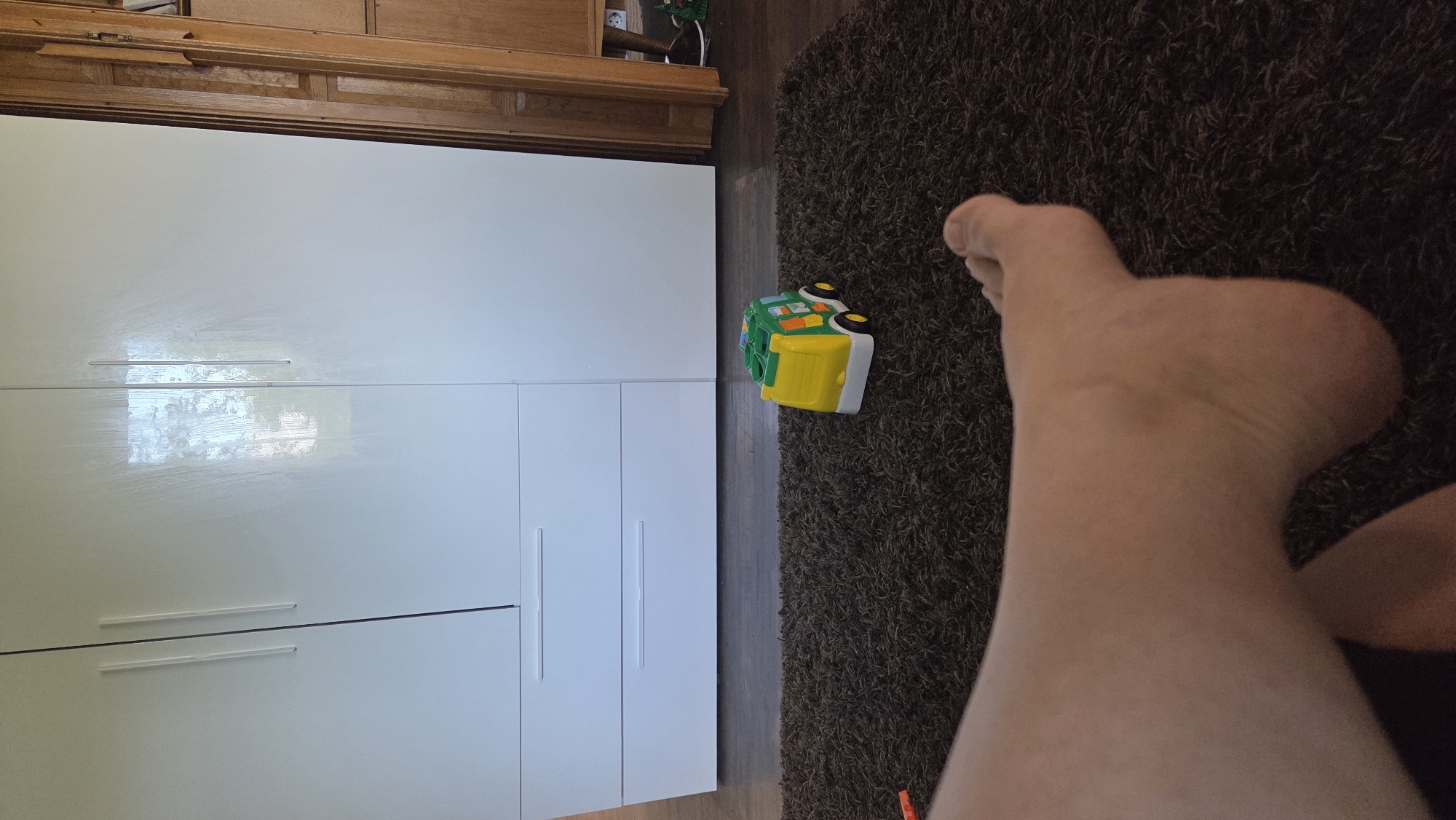 The image depicts an indoor scene, likely a bedroom or living room in a residence in Tynset, Norway. In the foreground, a person's bare foot is visible, resting on a dark brown shag carpet. To the right of the foot, a colorful toy garbage truck is placed on the carpet. In the background, a large, white, modern wardrobe with integrated handles dominates the scene. The wardrobe's doors have a glossy finish and reflect light, showing a blurred image of a window. To the right of the wardrobe, a wooden door frame is visible, and beyond it, a glimpse of another wooden door and wall suggests additional interior space. The lighting appears to be natural, coming from a window, and the overall atmosphere is casual and domestic.
