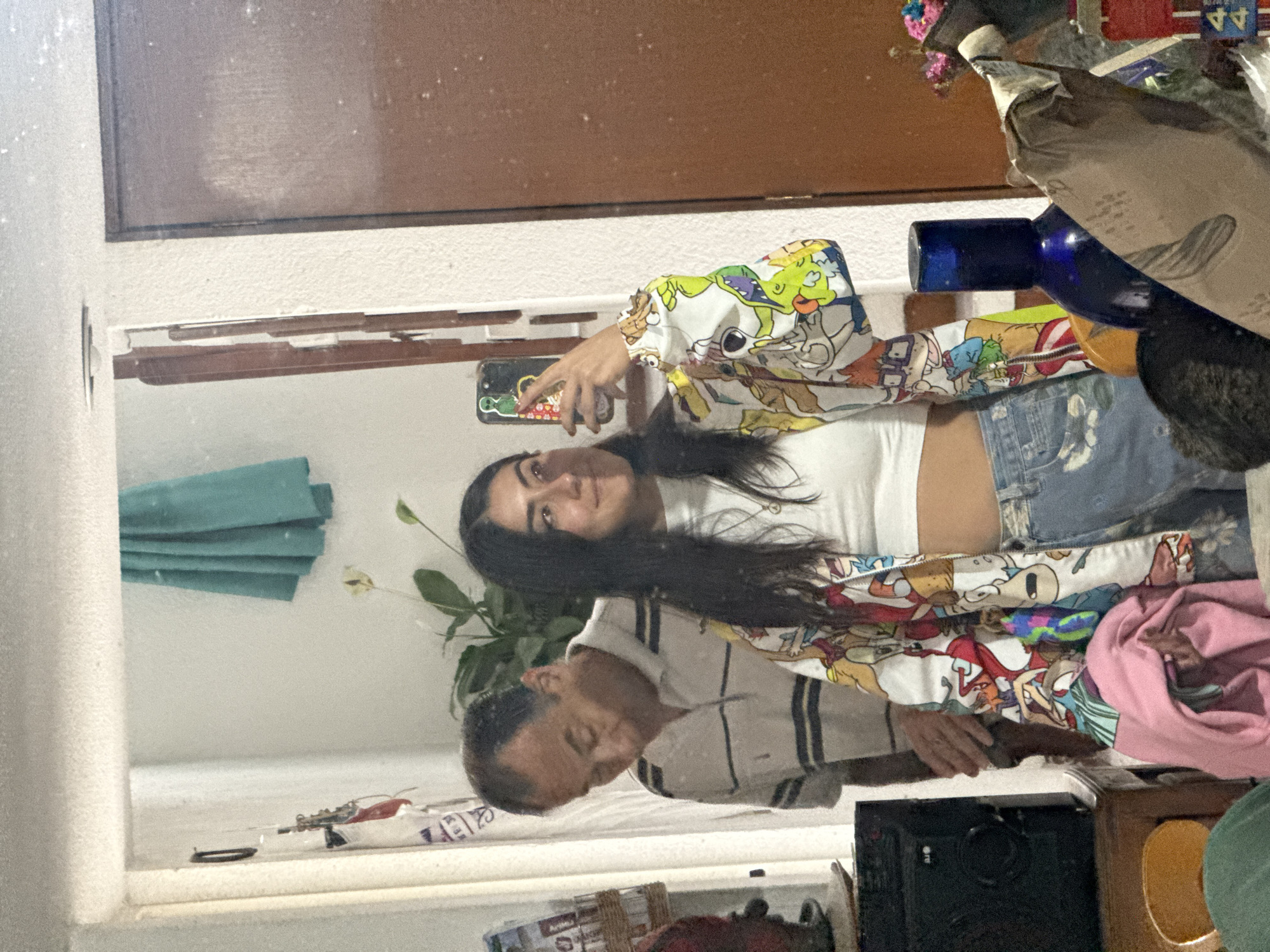 A woman is taking a selfie in a mirror. She is wearing a white top, denim shorts, and a jacket with cartoon characters. Behind her, in the reflection, a man is visible. The reflection suggests the setting is a domestic environment. The location is Puebla, Mexico, but there is no information from the image that can reveal which specific part of Puebla this scene is taken in. There is an assortment of objects visible, including a vase with a white flower, clothing, and various unidentified items on a shelf.