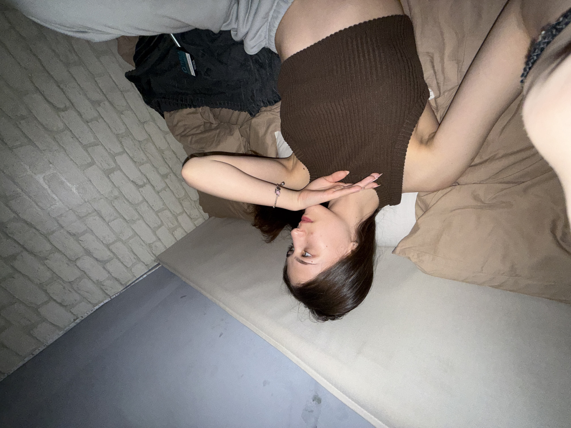 A young woman is lying on a bed, facing upwards. She is wearing a brown tank top and grey sweatpants. Her arms are raised, and her hands are near her face. The bed has beige bedding. A brick-patterned wall is visible in the background, along with a portion of the floor. Clothes are bundled on the bed near her waist. The image does not provide information to identify the specific location in Sofia, Bulgaria.