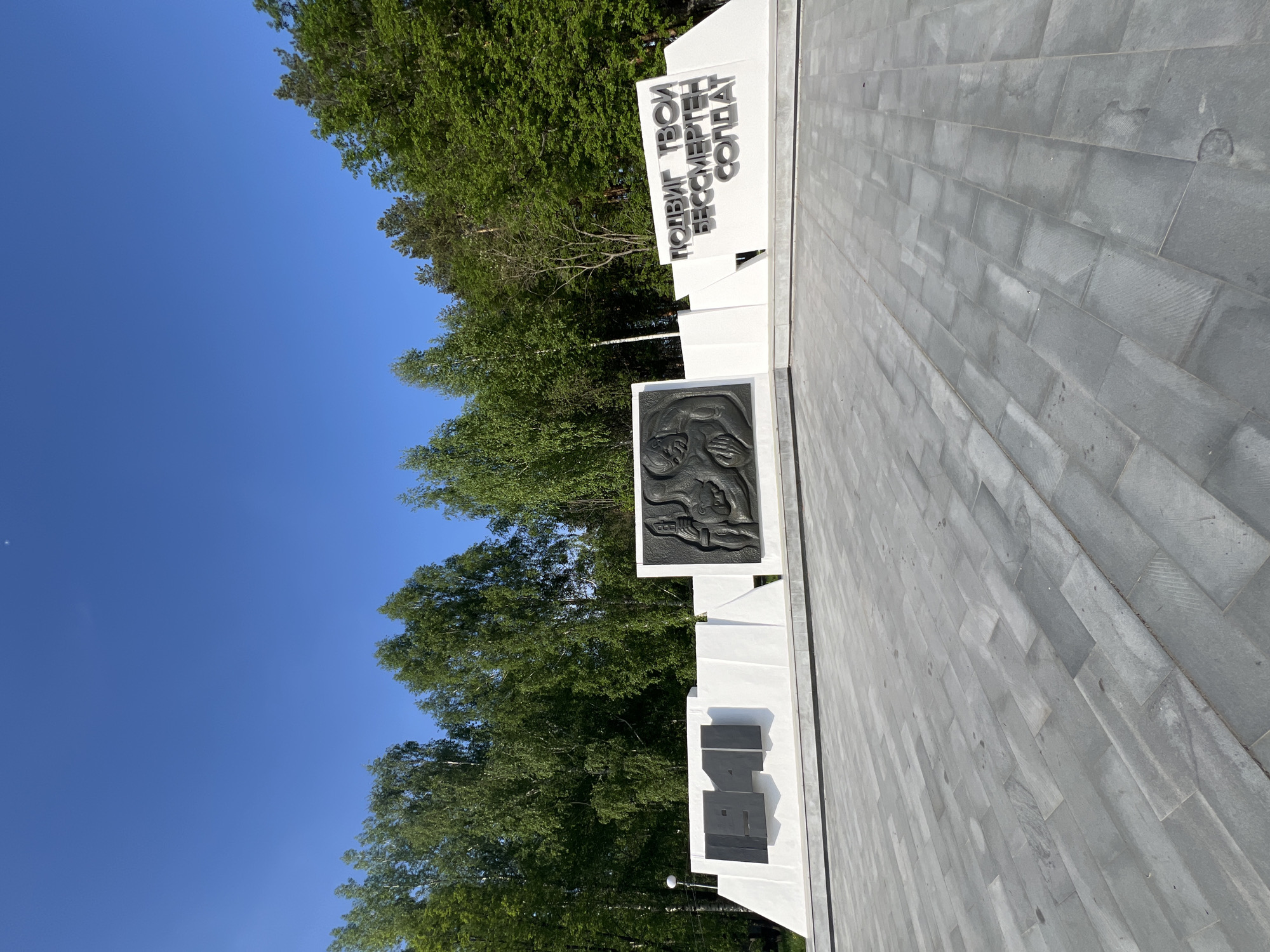 The image depicts a monument located in Chaikovsky, Russia. The monument is white with gray stone tiling leading up to it. The monument has a dark relief sculpture and Cyrillic text that reads "ПОДВИГ ТВОЙ БЕССМЕРТЕН СОЛДАТ". The monument also displays the year "1941" in dark gray, representing the start of the Great Patriotic War. The background shows green trees and a clear blue sky. The image is taken at ground level, looking up at the monument.