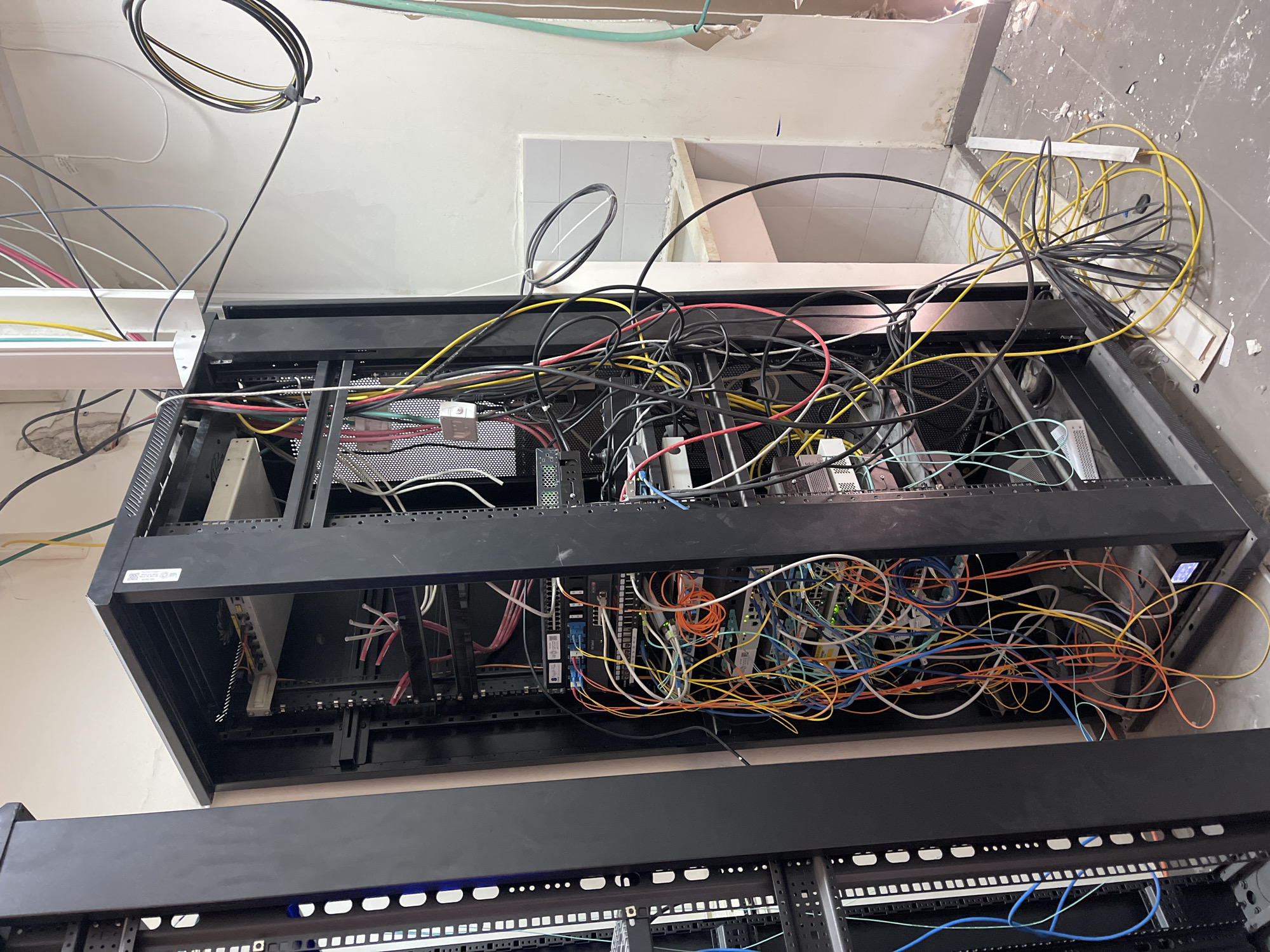A server rack is visible in the foreground of the image, filled with various network cables and equipment. The rack is black and appears to be made of metal. Cables of different colors, including red, orange, yellow, blue, and black, are neatly organized in some sections but appear tangled in others. Several server devices or switches are mounted within the rack. To the left of the server rack, another black rack is partially visible. In the background, a white wall with some visible damage and a tiled alcove are present. A sink basin is situated within the alcove. Electrical cords and cables are also seen running along the wall and ceiling. The floor is made of concrete tiles and has debris scattered across it, suggesting an ongoing construction or renovation project. The image does not contain any people or specific landmarks that would indicate its location in Tel-Aviv, Israel.