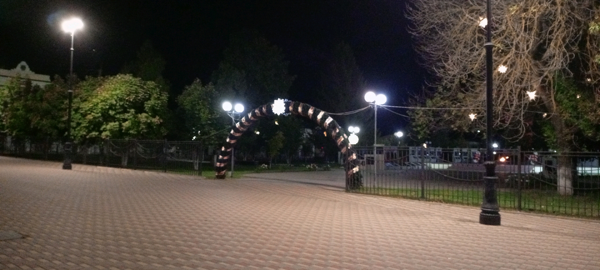 The image depicts a nighttime scene in the city of Kirsanov, Russia, likely a park or public square. In the foreground, a paved walkway with a brick-like pattern extends across the frame. To the left, a lamppost with a single bright light stands prominently. Further back, a large, arched structure decorated with black and white stripes and illuminated with lights spans across the scene. On the right side of the image, another lamppost is visible, adorned with small, star-shaped lights. The background is a dark expanse with trees and some fencing, suggesting a relatively open area. No people are visible in the image.