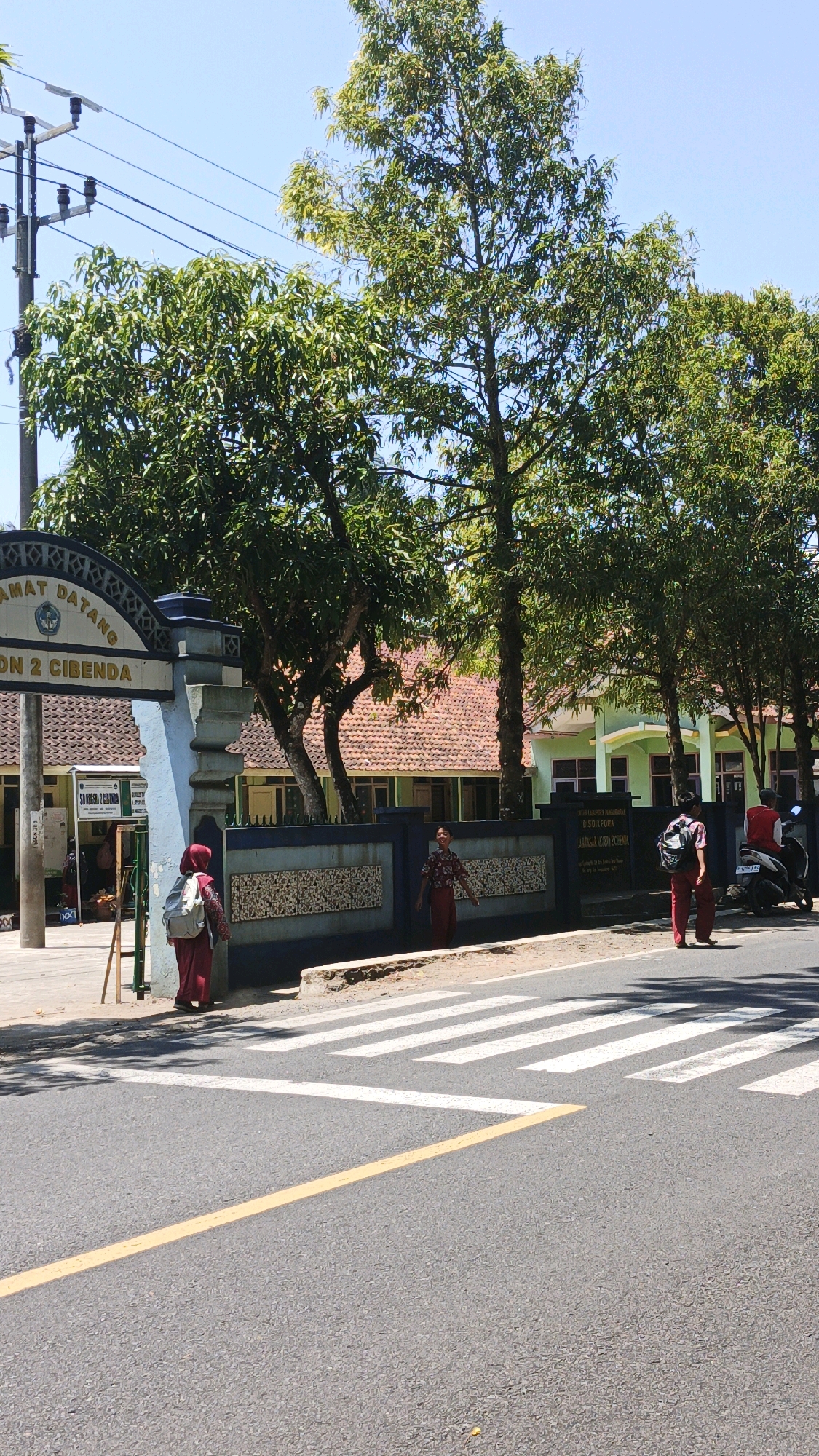 An exterior medium shot captures the entrance of Sekolah Dasar Negeri 2 Cibenda, a primary school in Cibenda, Indonesia, on a sunny day. The school's blue and white archway, adorned with the Indonesian national symbol, welcomes visitors with the text "SELAMAT DATANG" and "SDN 2 CIBENDA". A dark blue wall with ornate white patterns separates the school grounds from the street. A pedestrian crossing with white stripes is marked on the asphalt road in the foreground.
Three individuals are visible near the entrance. A young person wearing a red hijab and a light-colored backpack stands on the sidewalk facing away from the camera, entering the school grounds. To their right, a person in a red patterned shirt and pants stands near the wall, looking towards the camera with a slight smile. Further to the right, another person in red pants and a white shirt with a red vest walks away from the camera, carrying a backpack, and is near a parked motorcycle with a rider wearing a red shirt and white vest.
The school building, visible behind the wall, features orange tiled roofs and light green walls with dark framed windows. Lush green trees with dense foliage flank the entrance and canopy over the school grounds and parts of the street, casting dappled shadows. Power lines are strung across the bright blue sky, supported by utility poles on the left side of the frame. The overall scene depicts a typical day at an Indonesian primary school.