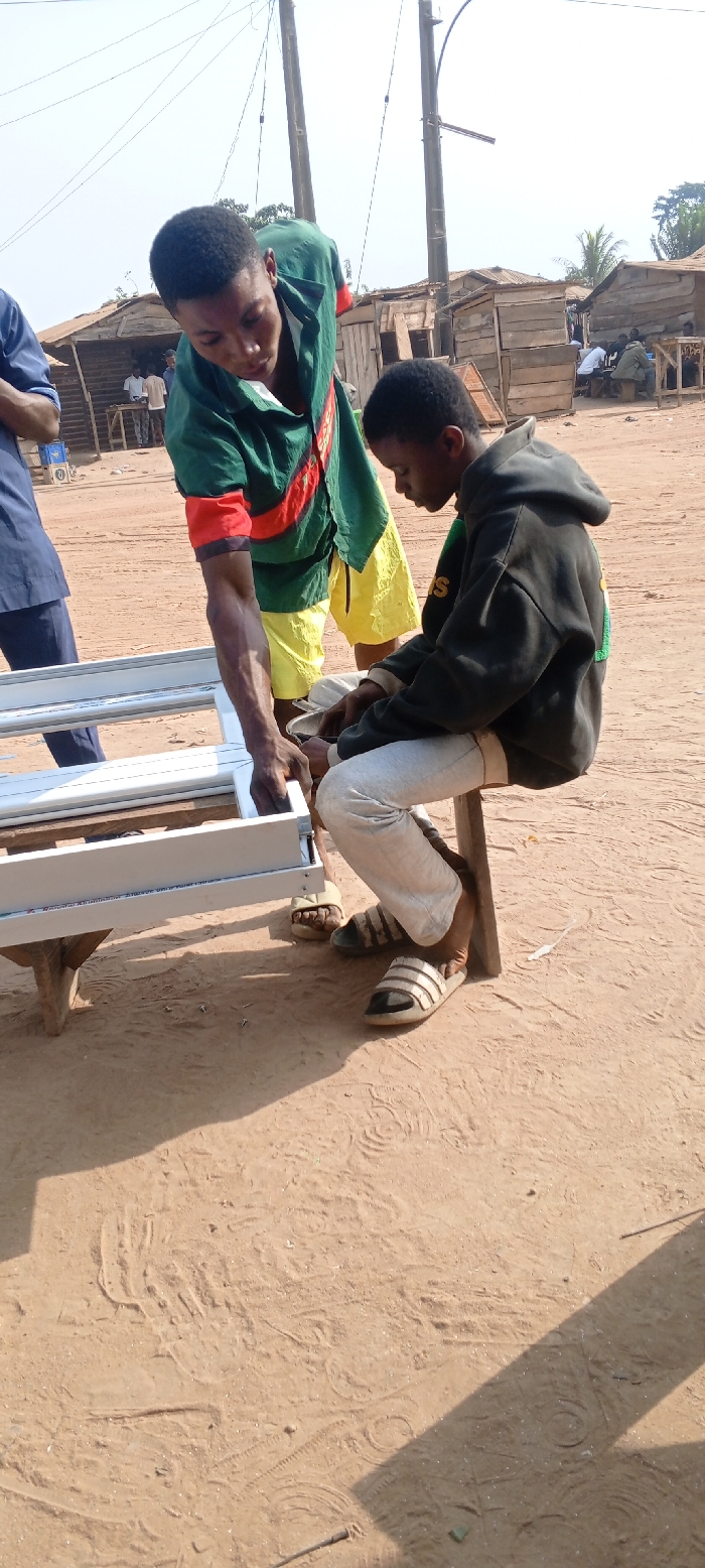 This image captures an outdoor scene in Ago-Abo, Nigeria, under direct sunlight, displaying two males engaged in an assembly task. In the foreground, a white, rectangular aluminum frame, resembling a window or door frame, rests on a rudimentary wooden table. The male on the left, standing, wears a green, yellow, and red striped t-shirt with yellow shorts; he is leaning over the frame, utilizing his right hand to manipulate a component. The male on the right is seated on a wooden stool, wearing a dark hooded sweatshirt and light grey sweatpants; he is intently focused downwards, appearing to work on a smaller object in his lap. The ground consists of reddish-brown, bare earth, marked with footprints and tire tracks. In the background, several informal wooden structures with corrugated roofs are visible, along with multiple indistinct individuals. Two wooden utility poles with overhead wires are prominent, and distant vegetation is visible against a clear sky.