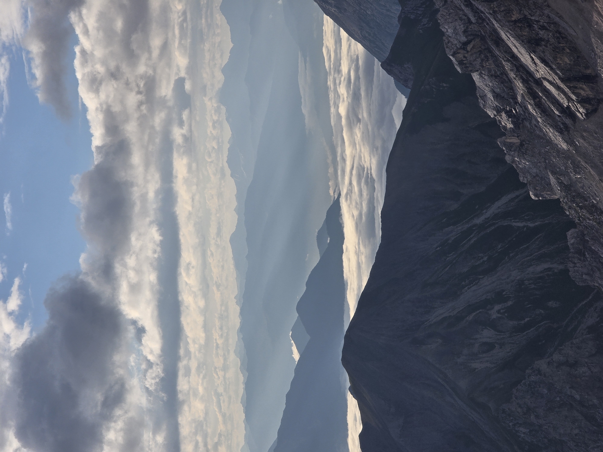 This is a vertical, full-shot photograph of a mountain range from a high vantage point. The foreground shows a steep, rocky slope in shadow, with a darker, heavily shadowed mountain peak dominating the lower left. In the midground, a sea of white clouds fills a valley, with lighter-toned mountain peaks emerging from the clouds. The background consists of multiple layers of hazy blue mountain ranges, fading into the distance. Above the mountains, the sky is a mix of blue and gray clouds, with beams of sunlight filtering through. The image depicts a dramatic mountain landscape under a cloudy sky. The exact location in Garmisch-Partenkirchen, Germany, is not specified.