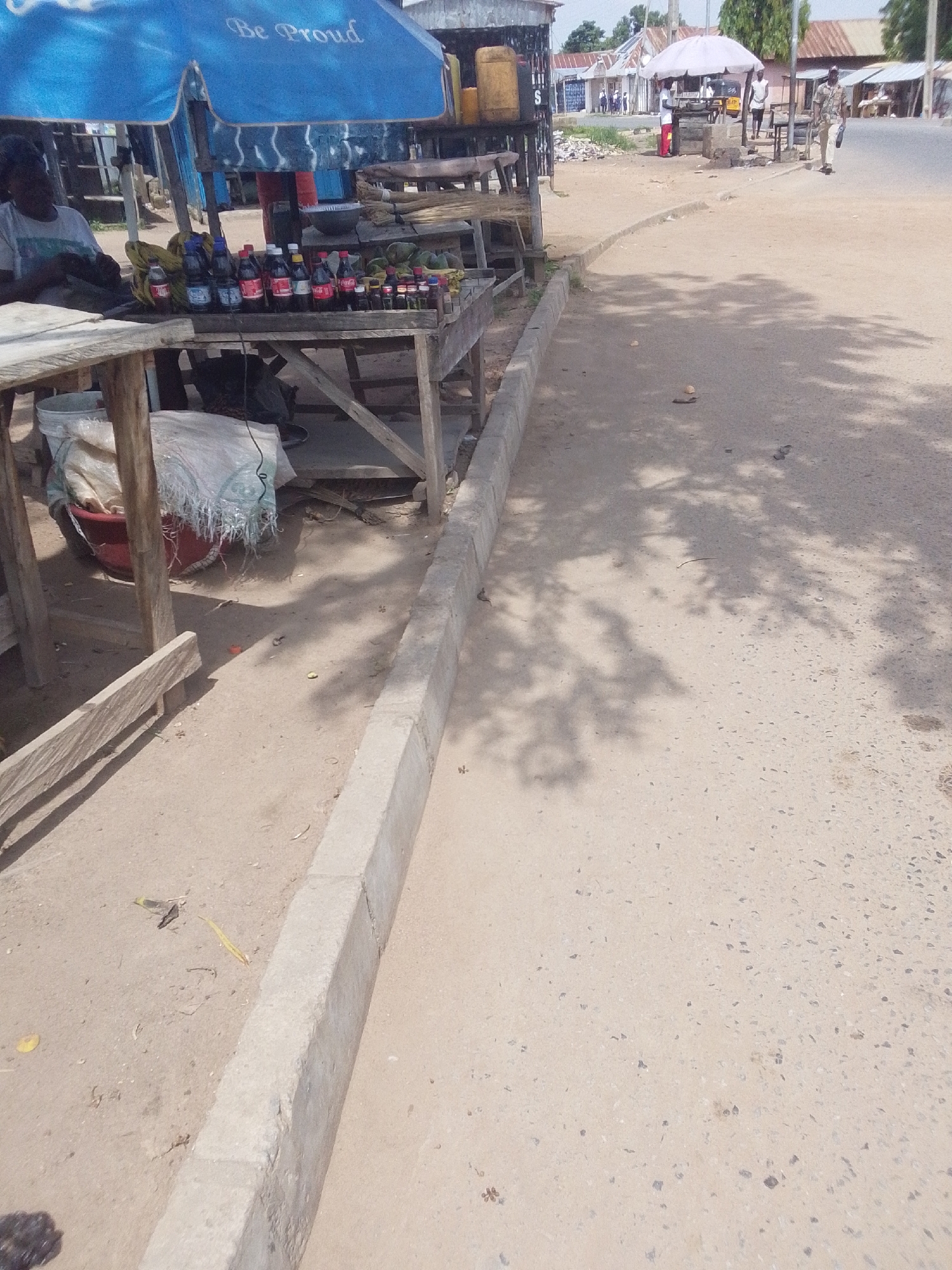 This image depicts a street scene in Jalingo, Nigeria, likely during daylight hours under a clear sky. The foreground is dominated by a dusty, uneven ground with a concrete curb running along its edge. Shadows of trees are cast across the ground, suggesting it's a sunny day.
In the mid-ground, a roadside stall is set up under a bright blue umbrella with the text "Be Proud" visible. The stall displays a variety of bottled drinks and small vials, likely for sale. A person, partially obscured by the umbrella, is seated behind the stall. To the left of the stall, a wooden table holds a red bucket filled with white and blue-ish bagged items.
Further into the background, across a paved road, more stalls and buildings can be seen. One stall has a white umbrella, and several people are present, some appearing to be vendors and customers. A man in a military-style uniform is walking on the right side of the road. The buildings have corrugated iron roofs, typical of the region. The overall impression is of a busy, informal marketplace situated along a street. The atmosphere appears to be one of commerce and daily life in a Nigerian town.
