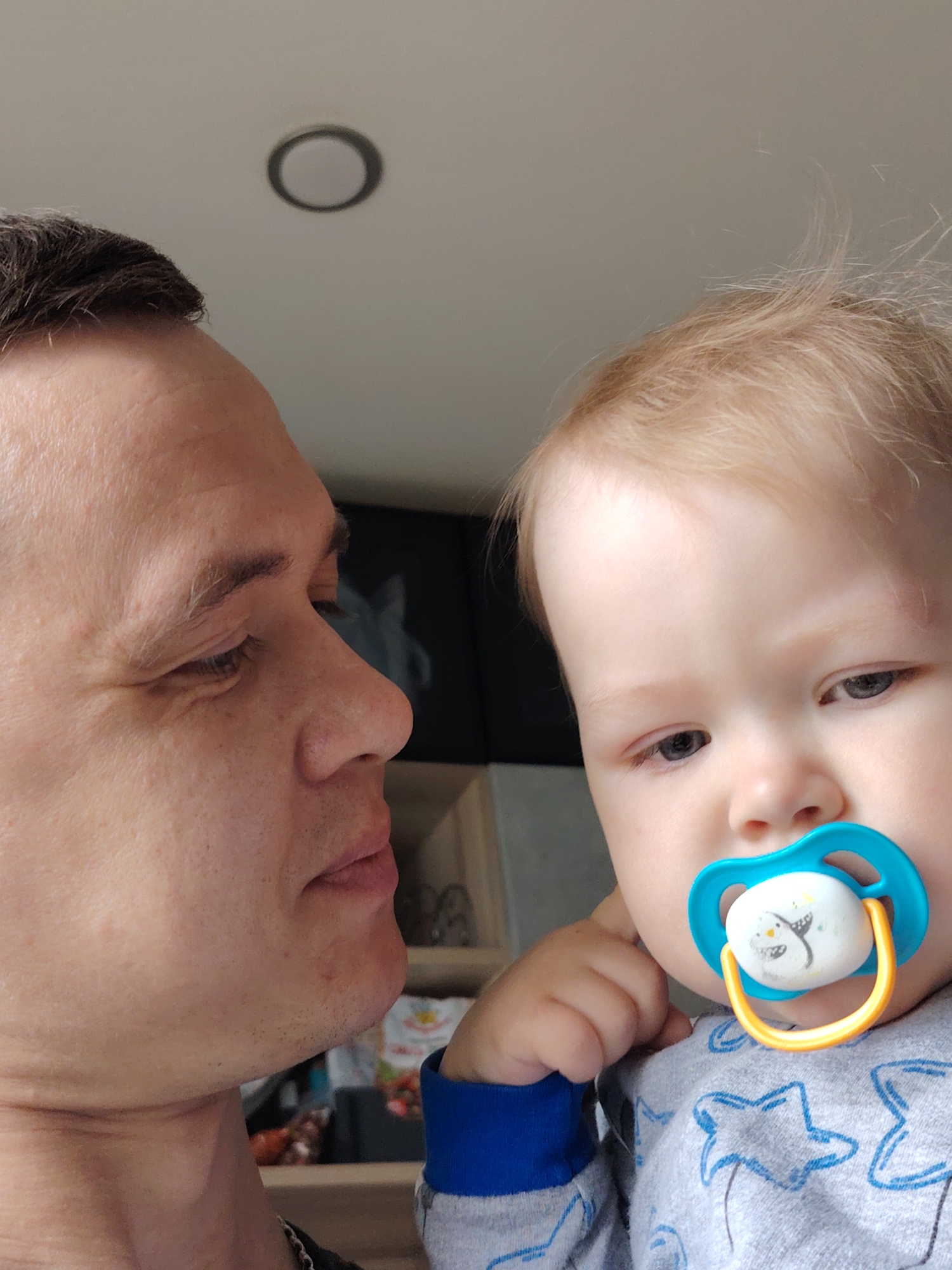 A medium shot captures a fair-skinned man and a baby. The man is on the left, turned slightly to the right, looking at the baby. He has short, dark hair and is wearing a dark shirt. The baby is on the right, facing forward with a blue and yellow pacifier in their mouth. They have fair skin and blond hair. The baby is wearing a gray long-sleeve shirt with blue star patterns. The background is a beige-colored wall with a circular light fixture near the top of the frame. A partially visible cabinet is behind the man, revealing various items, including what appears to be a package of strawberries. The location appears to be indoors, possibly a residence. There is no indication that the location is in Chaikovsky, Russia.