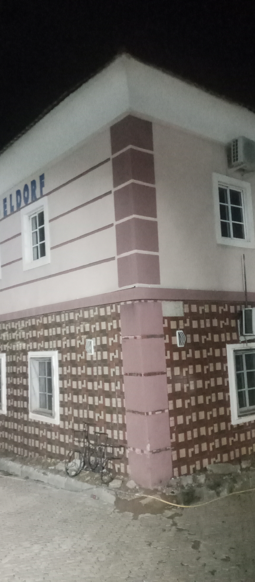 The image shows a building with the word "ELDORF" on its facade. The building has a light-colored exterior with pink accents, including decorative horizontal stripes and square tile patterns on the lower section. There are several windows visible, each with white frames and multiple panes. An air conditioning unit is mounted on the exterior wall near one of the windows. In the foreground, there is a paved area, possibly a driveway or courtyard, with a decorative metal bicycle-like structure. The scene appears to be at night or in low light conditions, as indicated by the dark sky.