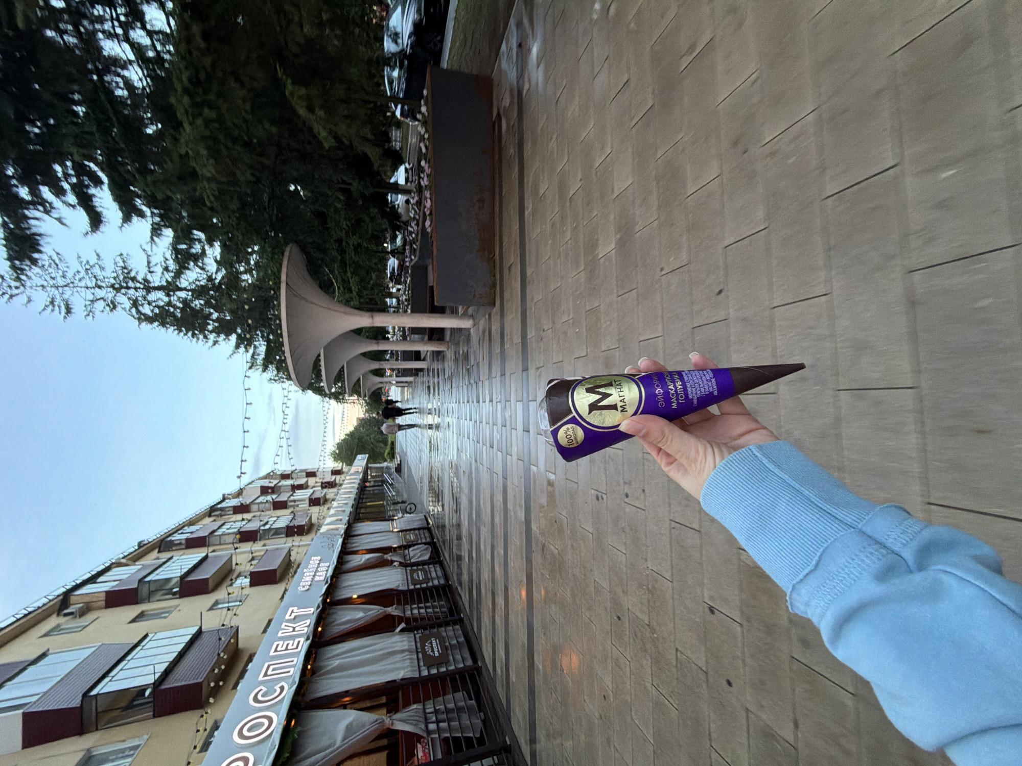 The image depicts a person's hand holding a Magnum ice cream cone in a city setting. The ice cream cone is dark purple with gold lettering and features the Magnum logo. The person is wearing a light blue sweatshirt. The background shows a paved walkway with distinct rectangular stones, and to the left, there is a multi-story building with balconies and awnings, likely housing shops or cafes. Across the walkway, there are modern white structures, possibly public art installations or architectural features, and large evergreen trees line the right side of the scene. The ground appears damp, suggesting recent rain. In the distance, several cars are visible parked along the street, and a few pedestrians are walking further down the path. The overall atmosphere suggests a city street or promenade in Tujmazy, Russia, during either early morning or late evening, given the soft lighting.