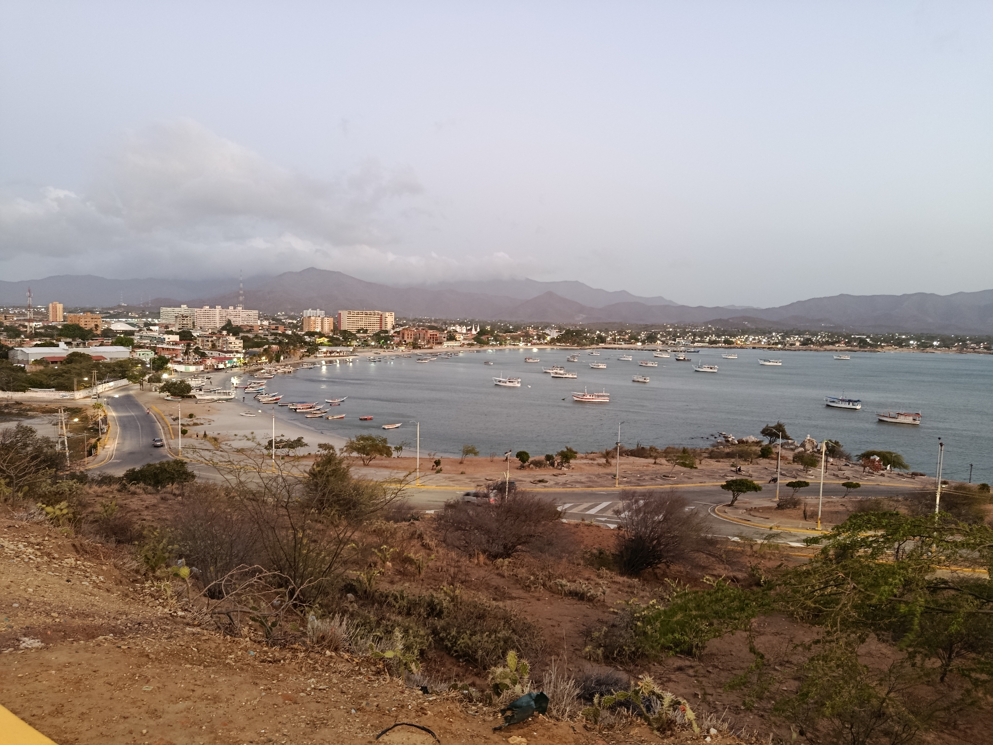 This image depicts a coastal vista in Juan Griego, Venezuela. The foreground is a dry, hilly terrain with sparse vegetation of shrubs and cacti. A road winds along the coastline, leading toward the town. The town itself is densely packed with buildings, including high-rise apartments, and extends along the bay. Numerous boats dot the bay's water, from small vessels near the beach to larger ones further out. In the background, mountains are shrouded in a cloudy sky. A car can be seen driving along the road by the beach. The overall lighting suggests either early morning or late afternoon.