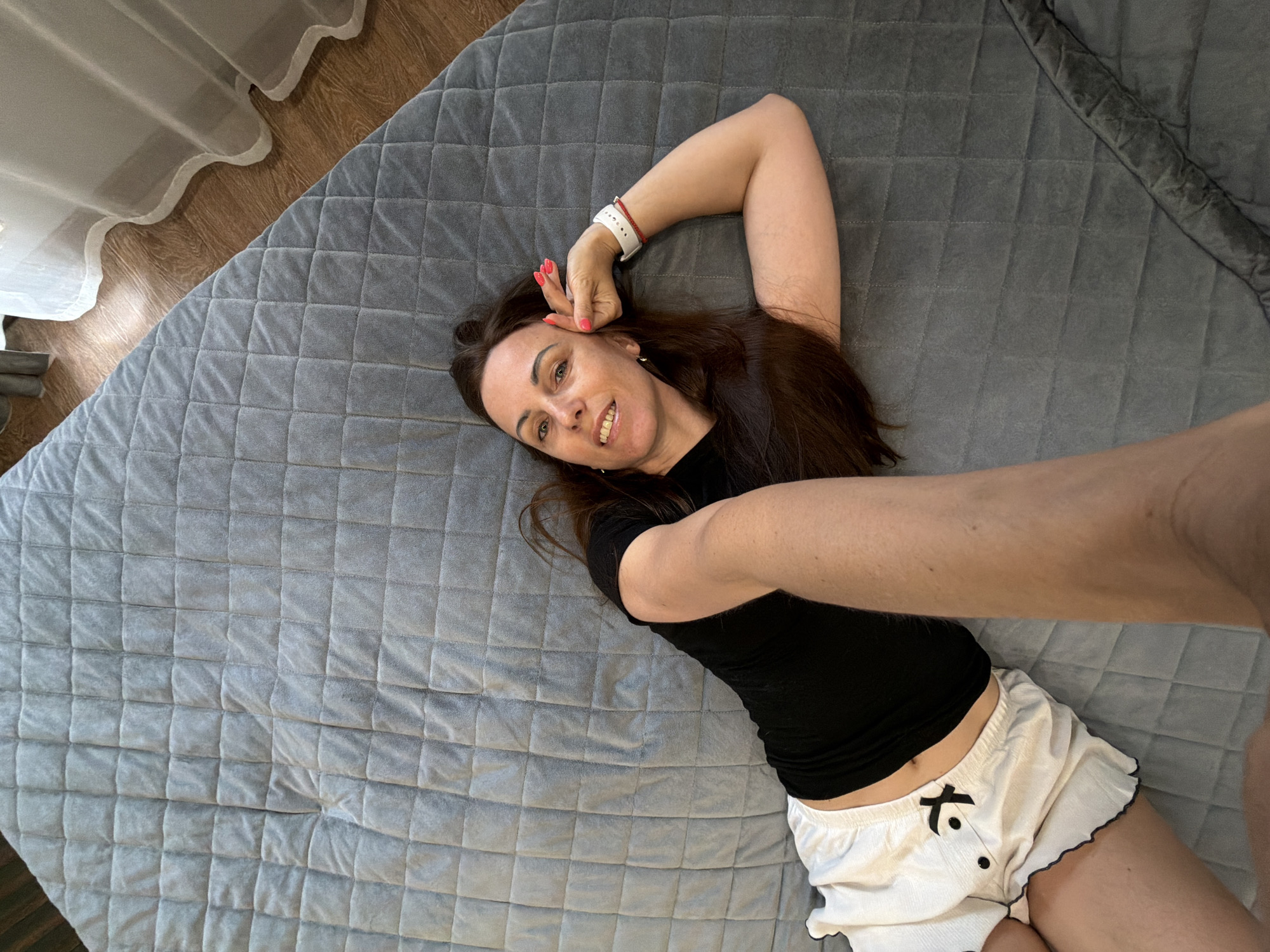An adult female is depicted lying on her back on a grey, square-quilted blanket or bedspread. Her head is positioned towards the bottom of the frame, and she is looking upwards towards the camera with a smile, revealing her teeth. She has long, dark brown hair spread around her head. She wears a black short-sleeved top and white shorts featuring a black bow accent on the front left. A white smartwatch or fitness tracker with a red band is visible on her left wrist (viewer's right). Her fingernails are painted a bright coral or pink color. Her right hand (viewer's left) is resting near her head, with fingers curled. A bare arm, presumably belonging to the person taking the photograph, extends downwards from the top center of the frame. A wooden floor and a white, sheer curtain are visible in the lower-left corner of the image. The environment appears to be an indoor setting. There are no visual cues within the image itself to confirm the specified location of Birobidzhan, Russia.