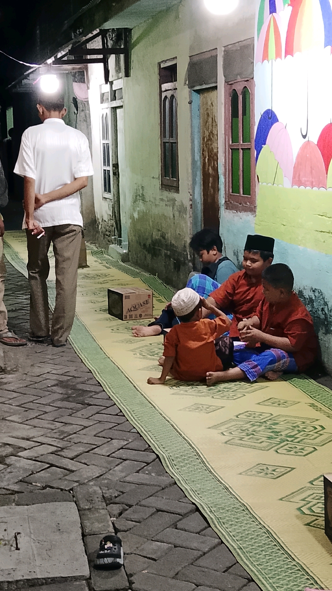 A group of four young boys are sitting on woven mats in a narrow alleyway in Randuagung, Indonesia. The boy on the far left is wearing a white cap and an orange shirt, looking down. Next to him is a boy in a red shirt and a black skullcap, interacting with the boy to his left. This boy is wearing a blue and red sarong and a red shirt. The fourth boy, on the far right, is wearing a blue checkered sarong and a blue shirt. He is leaning back and looking down. Behind them, a man in a white shirt and brown pants stands with his hands clasped behind his back, looking away from the camera. A box with "AQUASE" branding is placed on the mat in front of the boys. The alleyway has a rough brick pavement and a light-colored wall with windows and a painted mural of colorful umbrellas on the right side. A lamp hangs from the overhang of a building, illuminating the scene.