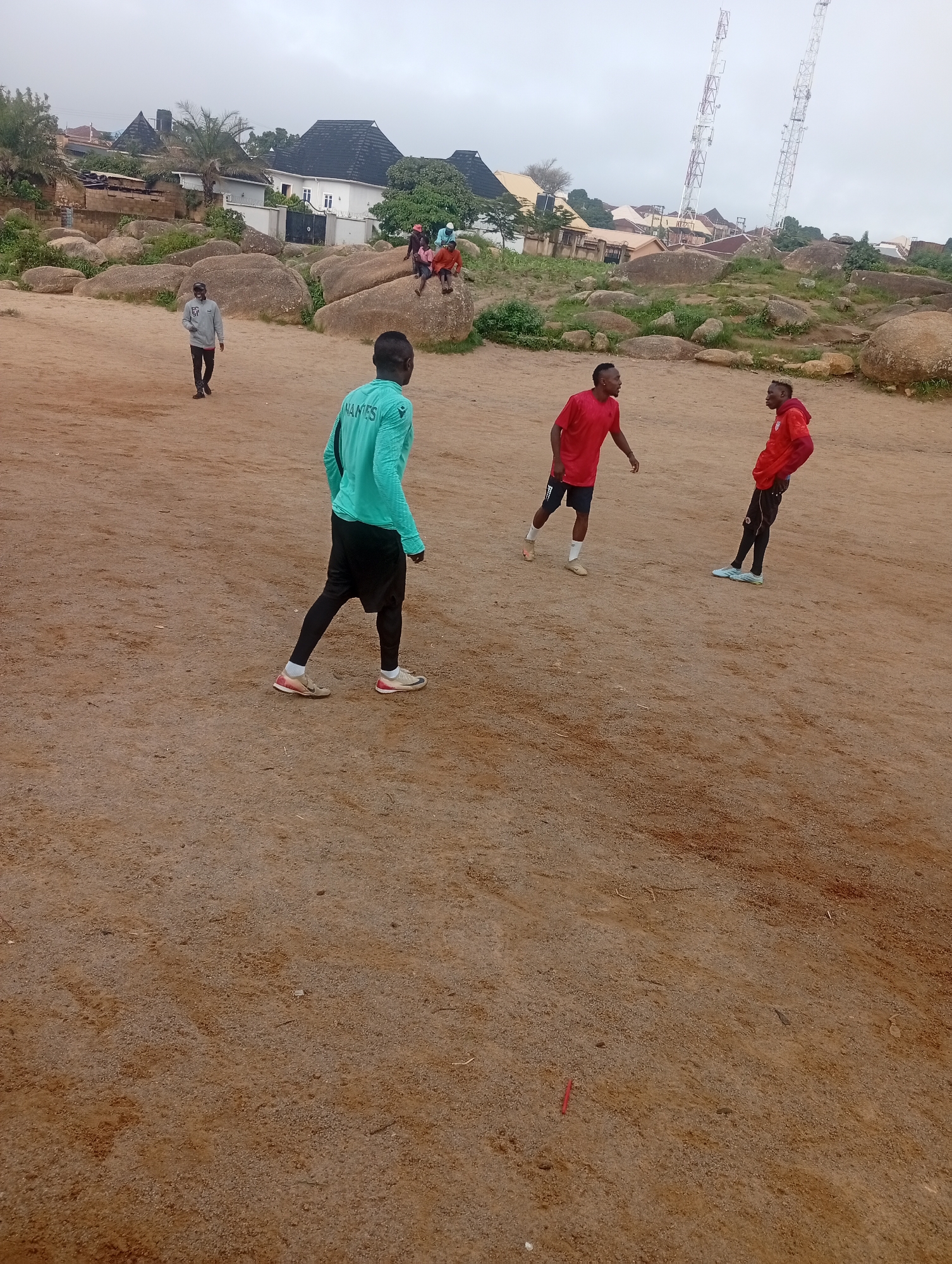 This image captures a scene in the city of Jos, Nigeria, likely during an overcast day. In the foreground, four young men are engaged in activity on a dirt field. One man, wearing a teal athletic shirt with "NAMES" printed on the back, black shorts, and black leggings, is facing away from the camera, walking. Two other men are wearing red shirts and dark shorts. One of them, on the right, is facing the camera and appears to be in motion, while the other on the left is facing away from the camera and also appears to be moving. A fourth man, further in the background and to the left, is wearing a grey sweatshirt and dark pants, walking. The background is dominated by large, weathered boulders and scattered green vegetation. In the distance, several buildings with dark roofs are visible, along with two tall communication towers. The overall impression is of a recreational area characterized by rocky terrain and a backdrop of urban development.