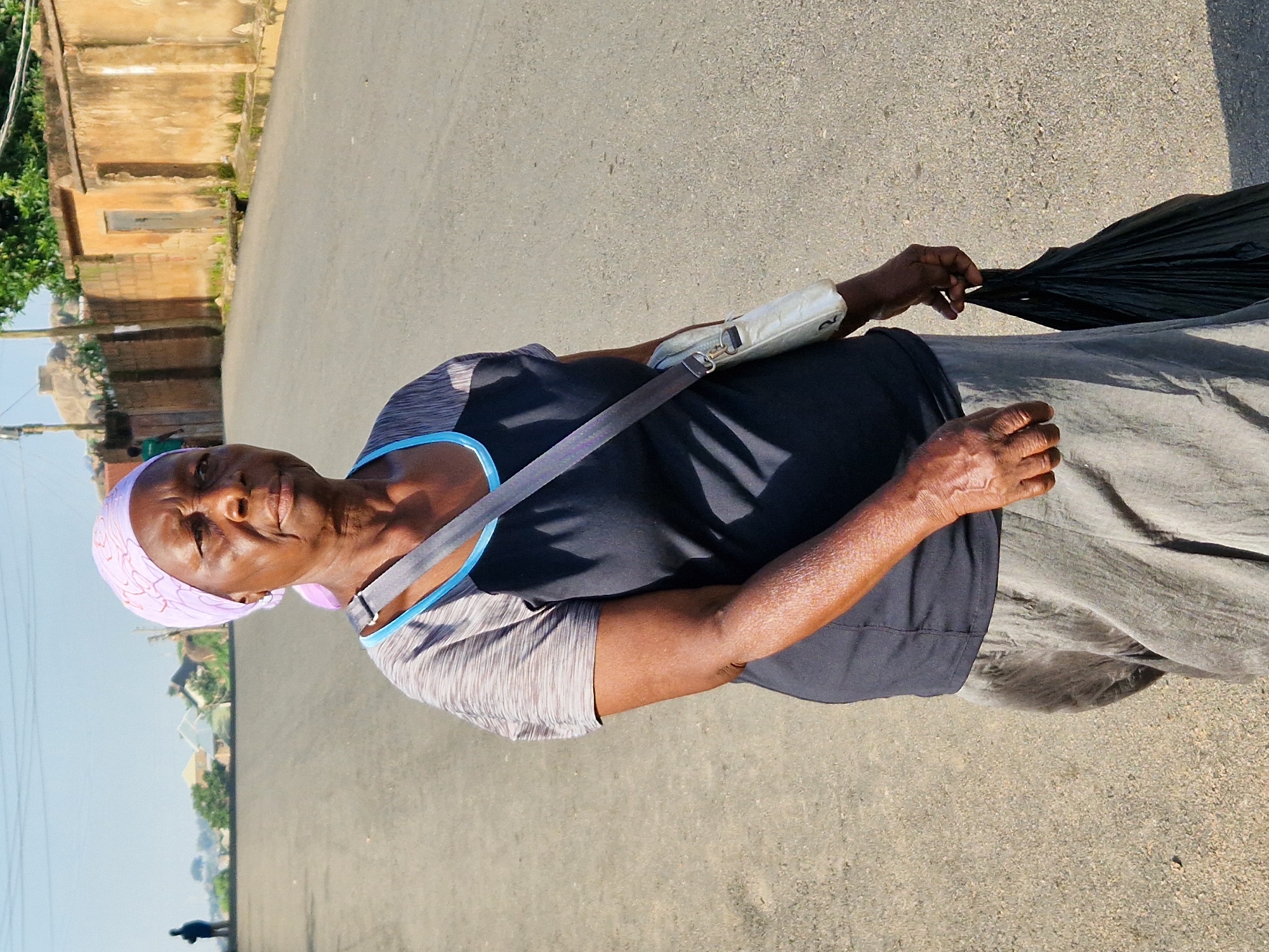 A dark-skinned woman wearing a light purple headscarf and a grey and blue shirt is walking down a paved road in an urban area. She has a light-colored crossbody bag slung over her shoulder and is carrying a black plastic bag in her right hand. The road is lined with buildings made of brick and concrete, some of which appear to be in disrepair. Trees and vegetation are visible in the background, along with utility poles and wires. The sky is clear and blue. The woman is looking to her right, and her expression is neutral.