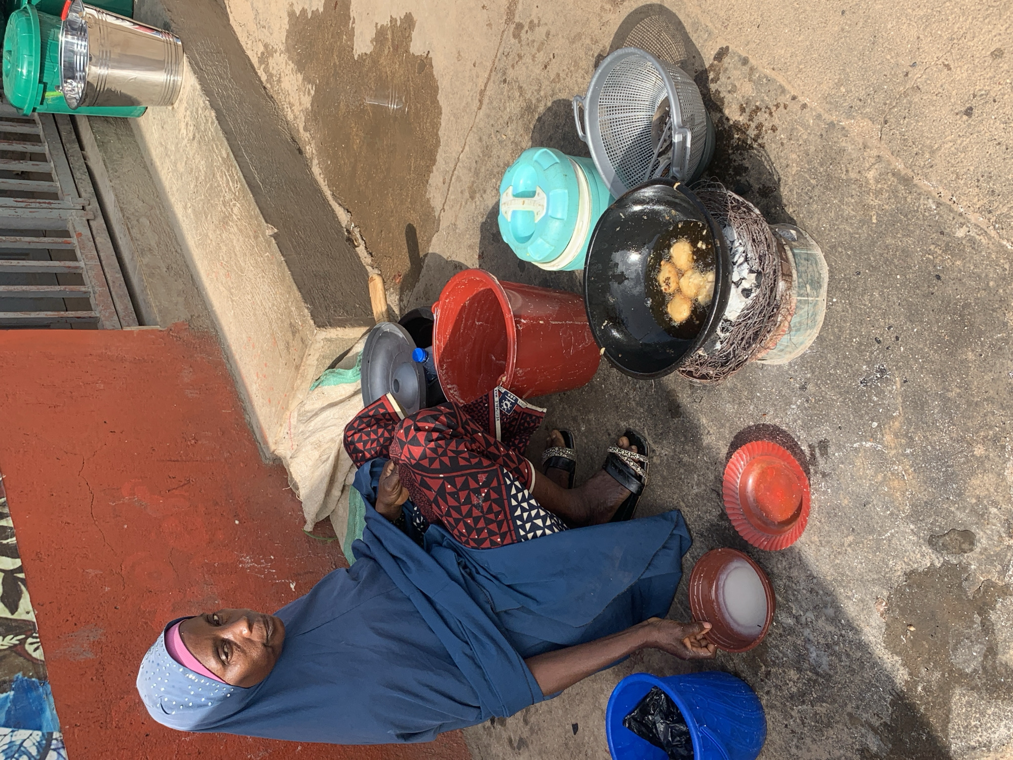 In Jos, Nigeria, a woman sits on the ground next to a red wall, her legs tucked beneath her. She wears a dark blue hijab and a patterned outfit. In front of her, a black wok sits on a makeshift stove, filled with oil and several golden-brown fried fritters. To her right, a grey mesh basket holds what appear to be more fritters, and next to it, a turquoise and white container. Further to the right, a metal bucket and two green bins are visible near a metal gate and concrete steps. A blue bucket filled with a black plastic bag sits to her left, beside a small brown bowl containing a white liquid, and a red plate. Water pools on the ground around the scene, reflecting the sunlight.