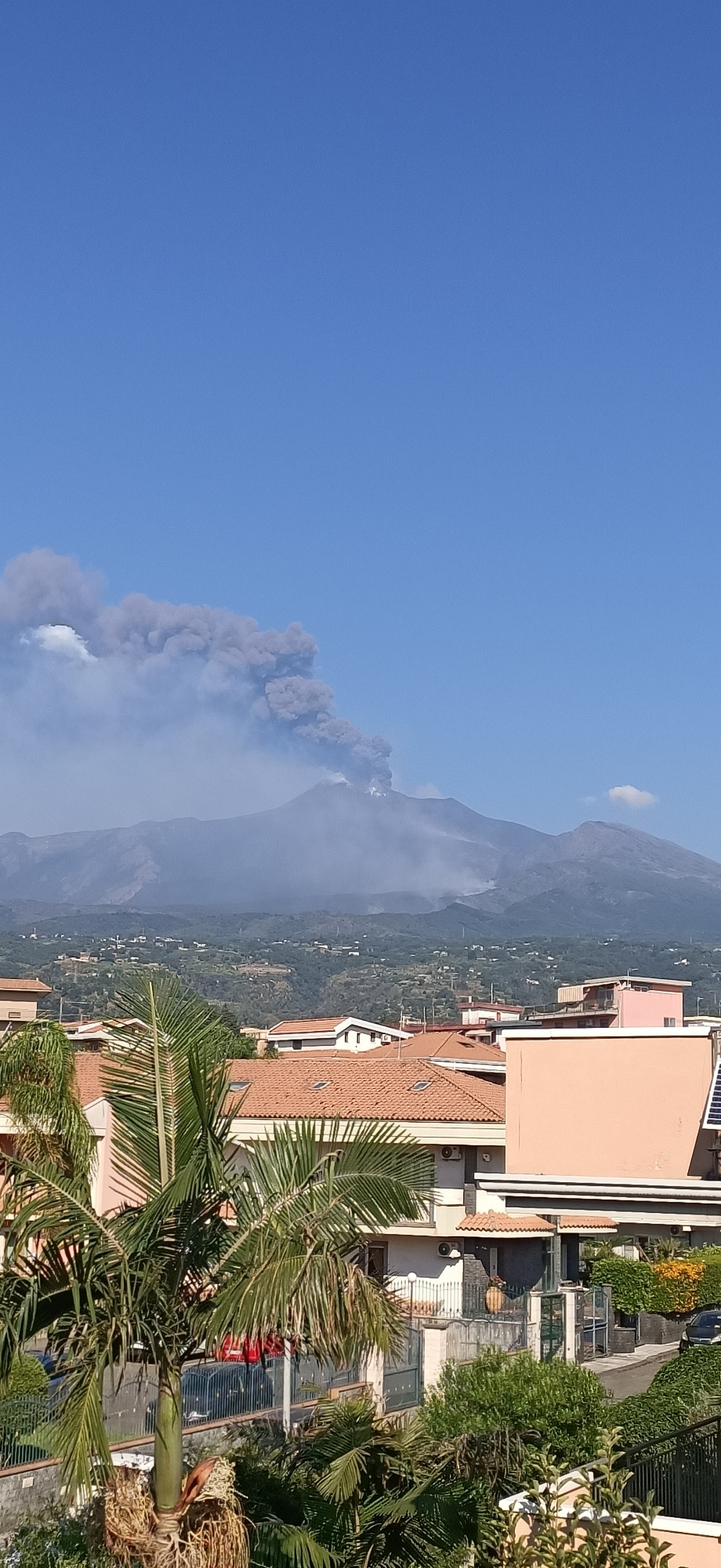 A wide shot captures a volcanic eruption against a clear blue sky. The volcano, a large mountain range in the background, is spewing a thick plume of dark gray smoke and ash high into the air. The smoke cloud is dense and billows outwards, obscuring the upper parts of the mountain. In the foreground, a suburban landscape is visible, with terracotta-tiled roofs of houses and lush green palm trees. The buildings are modern and appear to be residential. The overall scene depicts a natural event unfolding in a populated area.