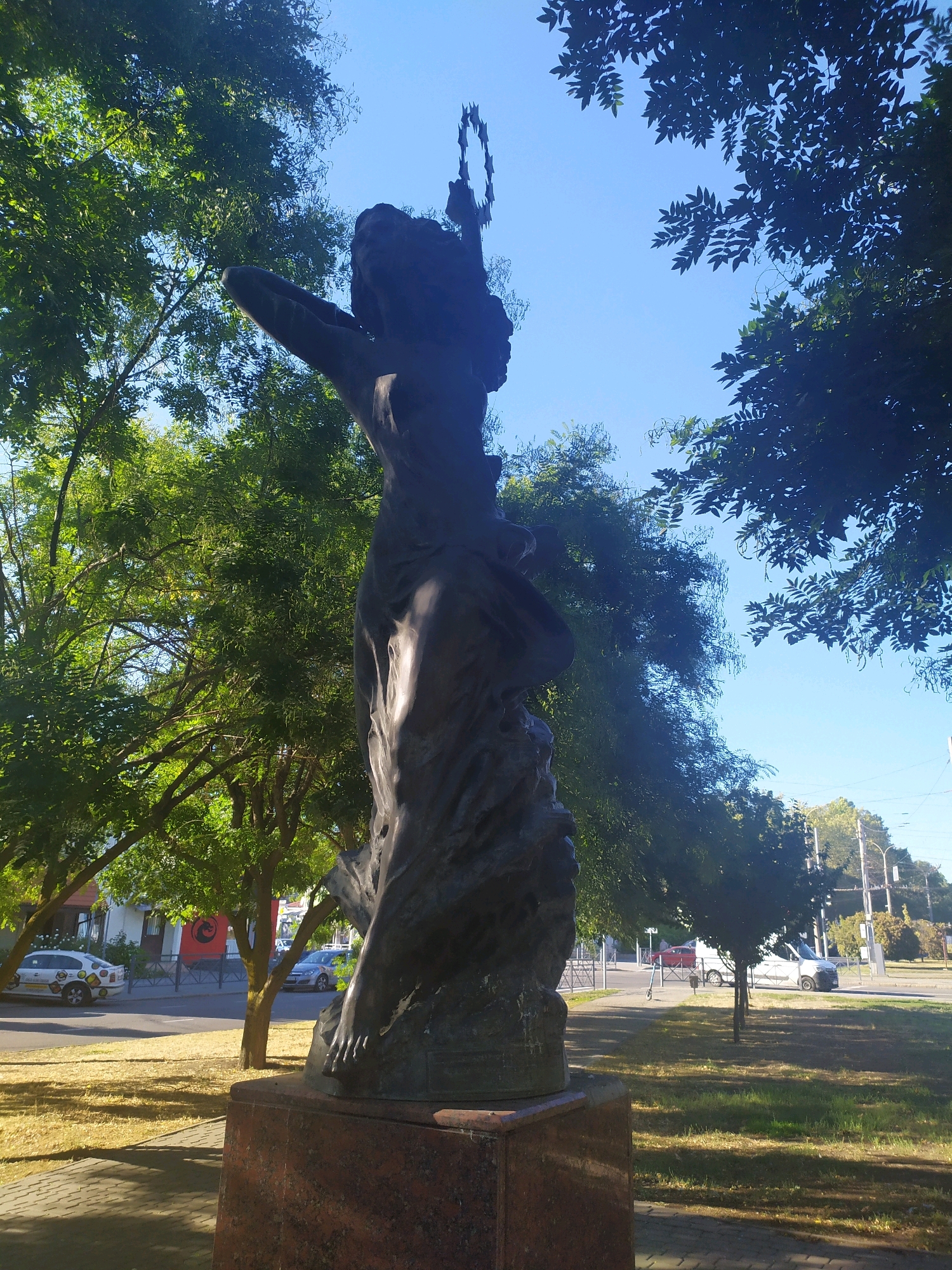 The photograph depicts a large, dark-colored statue of a human figure from a low angle, set against a clear blue sky and framed by green trees. The statue appears to be made of bronze or a similar metal, and its surface is textured and weathered. The figure is in a dynamic pose, with one arm extended upwards and the other bent at the elbow. The head of the statue is crowned with a circular, abstract element. The statue stands on a multi-layered pedestal, the bottom layer of which appears to be made of red granite.
In the background, a city street is visible with parked cars and trees lining the sidewalk. The scene suggests an urban park or a public square. The lighting indicates it is daytime, with sunlight casting shadows.
This statue is located in the city of Mykolaiv, Ukraine, and is known as the Monument to the Founders of Mykolaiv. It is situated on Soborna Square, near the Mykolaiv Regional Art Museum. The monument was erected in 2001 and is a significant landmark in the city.