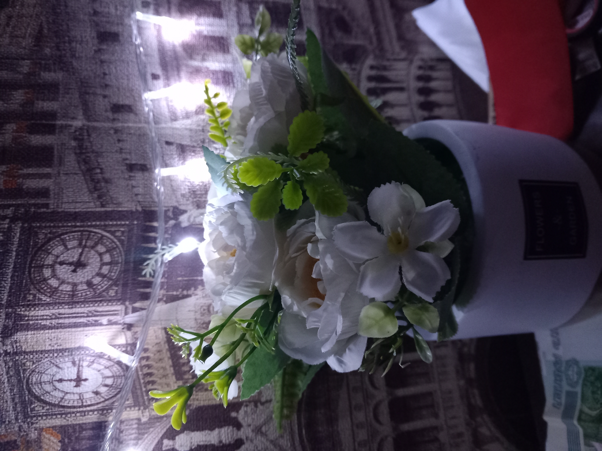 A bouquet of white artificial flowers and green foliage is displayed in a white pot. The pot has a label on the front that reads "FLOWERS & GARDEN". In the background, there is a tapestry or wallpaper depicting clock towers and architectural elements, possibly in London. String lights with white bulbs are draped across the scene, casting a warm glow. To the right of the pot, a red object and a white tissue box are visible. In the lower left corner, a portion of what appears to be currency is visible. The scene is lit by the string lights and possibly an overhead light source.