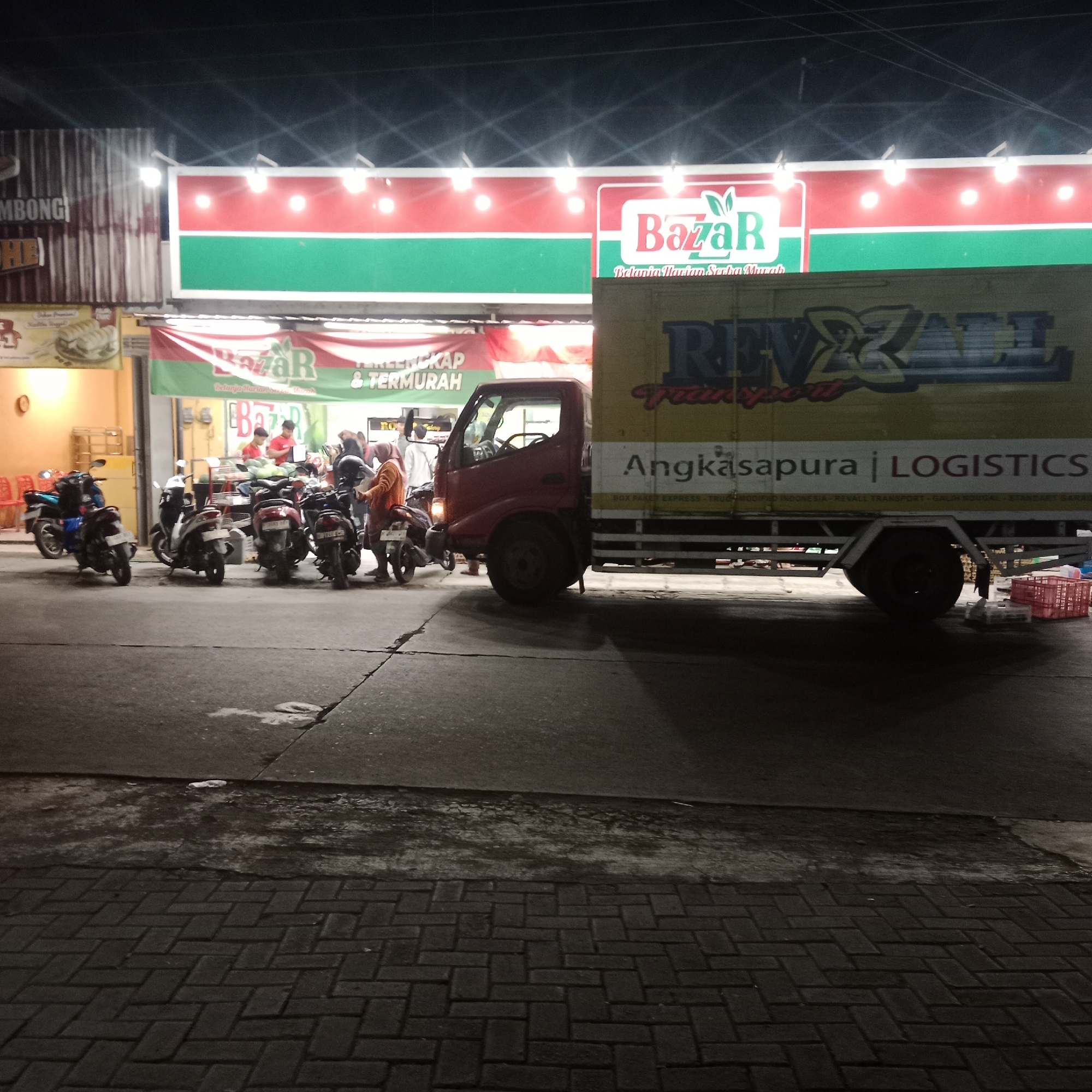 The image depicts a street scene at night in Gubug, Indonesia, in front of a BazaR store. A red truck with "ANGKASAPURA | LOGISTICS" and "REVAL TRANSPORT" written on its side is parked adjacent to the store. Several motorcycles are parked in front of the store, and a few people are visible near the entrance, some of whom appear to be employees. The store has a prominent red and green sign that reads "BazaR" and "Belanja Harian Serba Murah" (meaning "Daily Shopping for Everything Cheap"). The ground in front of the store is paved with interlocking bricks. The lighting suggests it is nighttime, with artificial lights illuminating the store and the street.