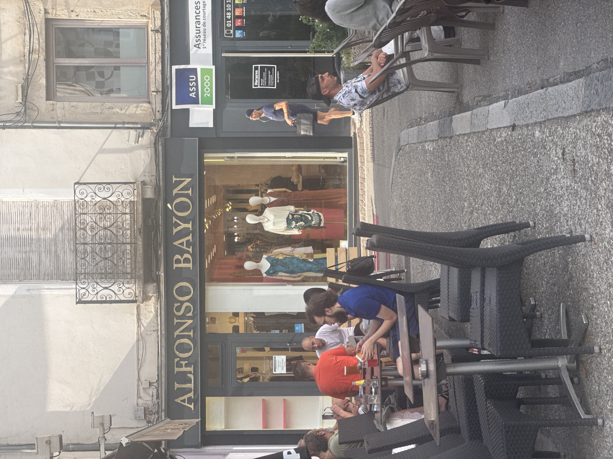The image depicts an outdoor seating area in front of a clothing store named "ALFONSO BAYÓN" on a sunny day. The store's facade features a large glass window displaying mannequins in dresses and racks of clothing. To the right of the clothing store, there is another establishment with a sign that reads "ASSU 2000" and "Assurances."
Several people are seated at tables in the foreground, appearing to be patrons of a cafe or restaurant. One man in an orange t-shirt and another in a blue t-shirt are visible, along with others whose faces are partially obscured. Drinks and food items are present on the tables.
A woman in a blue shirt and shorts is walking past the storefronts carrying a black bag and wearing headphones. The ground is paved with asphalt, and there are several dark wicker chairs and metal tables set up for outdoor dining.
The architecture of the buildings suggests a European urban setting, and the presence of "Vienne, France" in the prompt implies the location. The overall scene conveys a relaxed, everyday atmosphere of people enjoying leisure time in a commercial district.