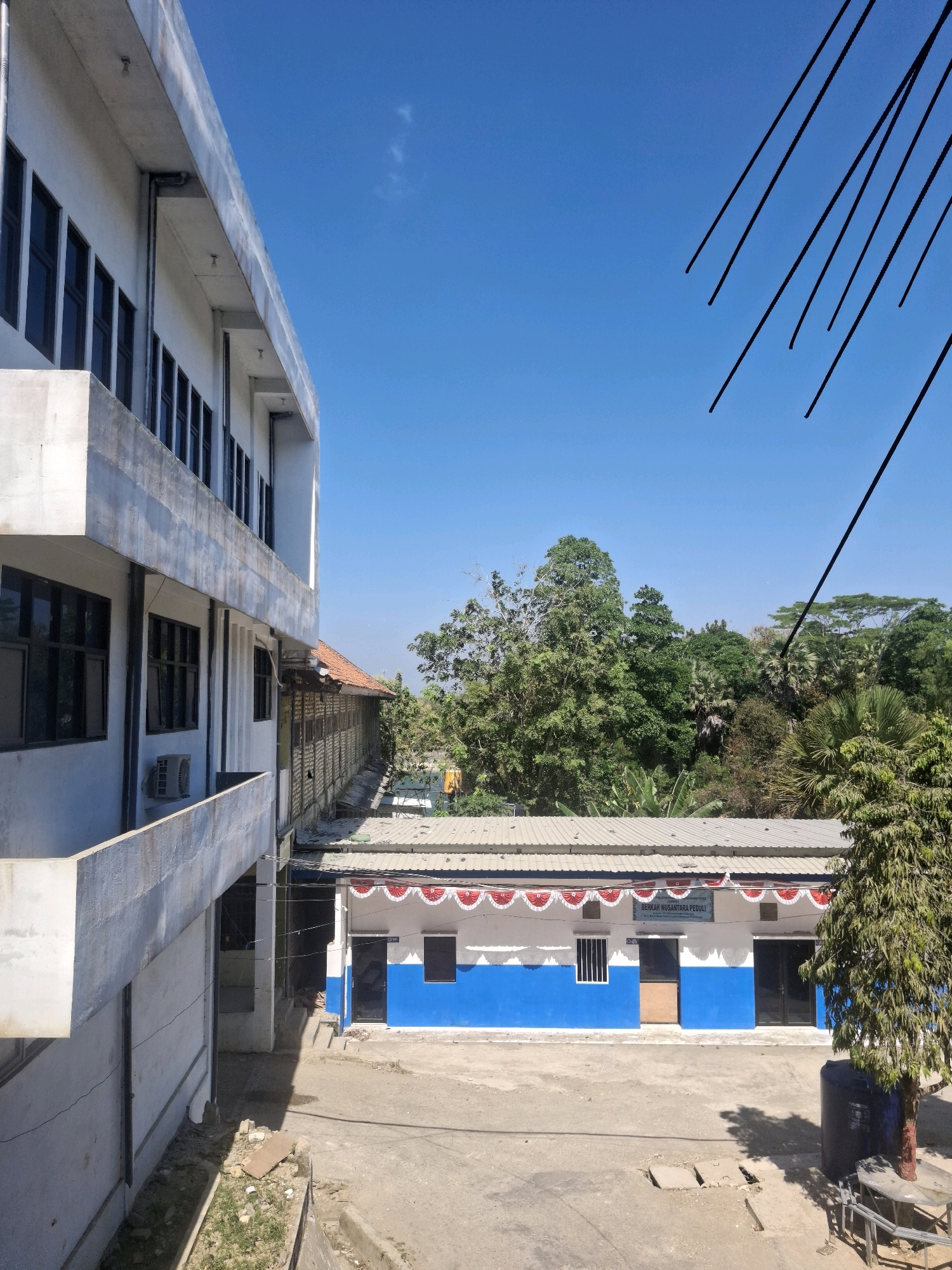The image depicts a view of a building complex under a clear blue sky. On the left is a multi-story white building with rows of dark-framed windows. Below the windows, an air conditioning unit is visible. To the right of this building, a lower, single-story building features a blue and white facade decorated with red and white bunting. This building has several doors and windows, one of which is a small window with white bars. Above this building, lush green trees form a dense canopy. In the foreground, a concrete ground surface is visible, with some debris, including a piece of plywood and some scattered stones. A large dark blue water tank stands near the single-story building, next to a small tree. The scene appears to be an outdoor area within a community or institutional setting. The location is Potoan Laok, Indonesia.