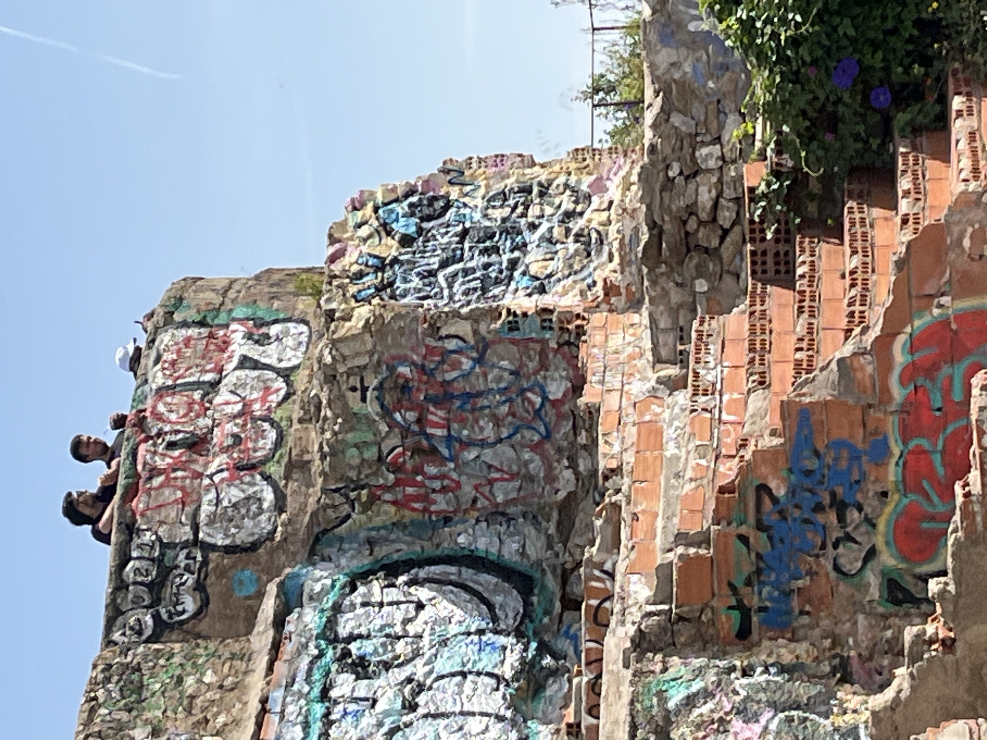 This media file captures a low-angle view of a crumbling, heavily graffitied structure against a bright, clear blue sky in Barcelona, Spain. The structure appears to be an old wall or ruin, constructed from a mix of rough stone blocks and red bricks, many of which are broken or irregularly placed, indicating disrepair.
On the far left, two individuals, likely men, are partially visible, peeking over the top edge of a graffiti-covered section of the wall. They are seen from the chest up, looking downwards and towards the right, appearing to observe something below or in front of the structure. One wears a dark shirt, the other a lighter one.
The entire visible surface of the stone and brickwork is covered in vibrant spray-painted graffiti. On the section where the people are, large, stylized white letters with red outlines are prominent. The central and right sections display a dense overlay of tags and bubble letters in various colors, including blue, red, white, and black, making specific words largely illegible. The graffiti forms abstract patterns rather than clear messages.
On the upper right side of the structure, green leafy plants, possibly vines, grow over the bricks, and a few purple, bell-shaped flowers, resembling Morning Glories, are visible. The sky in the upper left is clear blue, with a faint white contrail from an aircraft just discernible. The bright, even lighting suggests it is midday or early afternoon on a sunny day. The scene conveys an atmosphere typical of an urban environment with a strong presence of street art.
