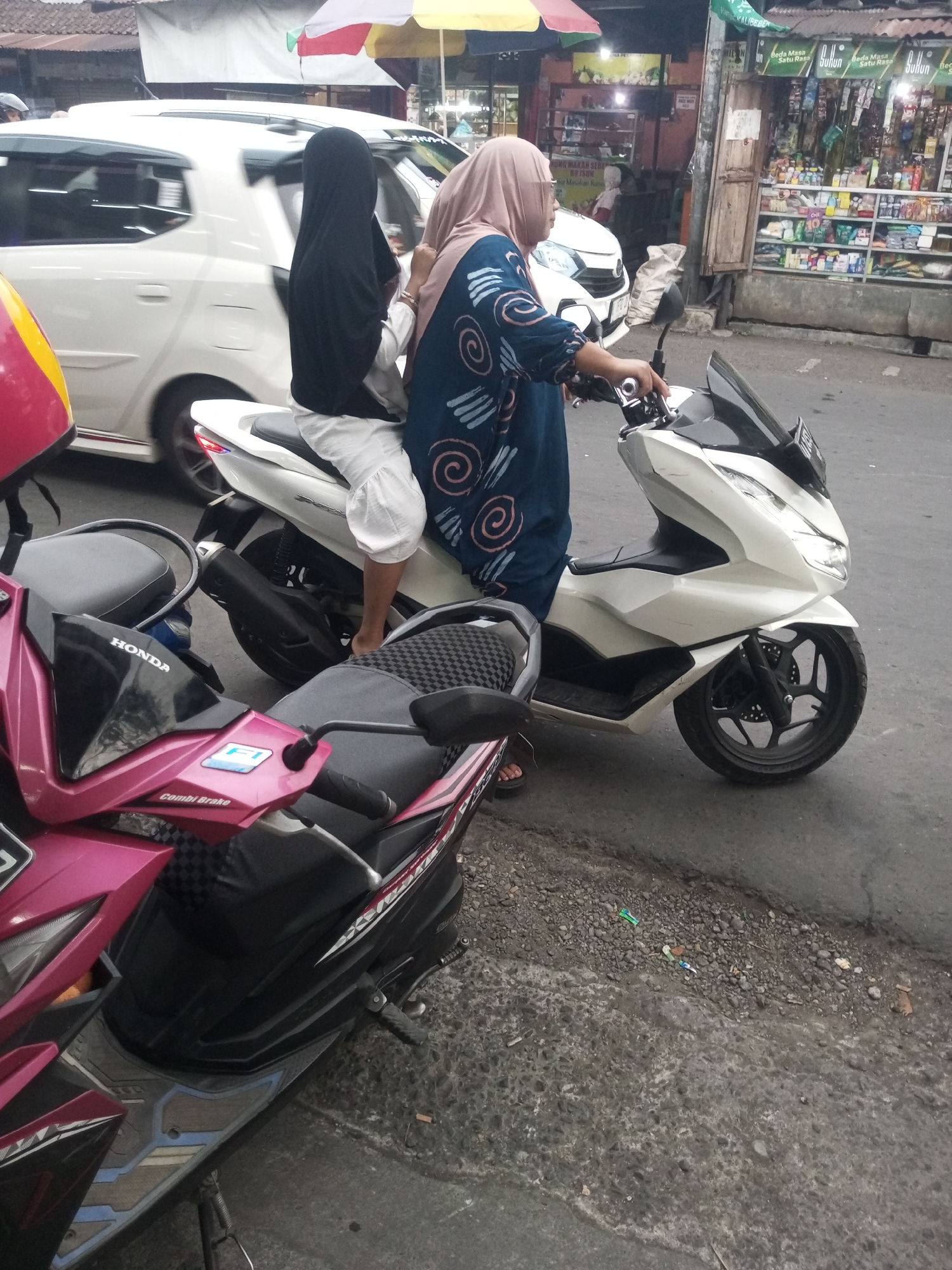 Two women are on a white scooter in a street. The woman in front is wearing a blue patterned dress and a light pink hijab. She is holding the handlebars and looking forward. The woman behind her is wearing a black hijab and a white shirt. She is sitting behind the driver. The scooter is parked on the side of a street. In the background, there are shops and a white car. The scene appears to be in Kalibeber, Indonesia, based on the signs visible in the background. The ground is paved with asphalt and there are some pebbles and dirt on the side of the road.