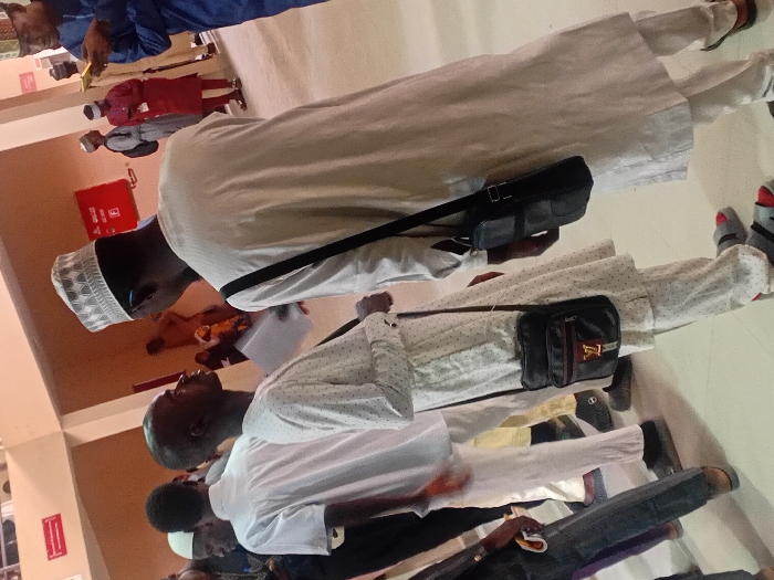 This image captures an indoor scene in the city of Kwazailewa, Nigeria, depicting a gathering of individuals within a modern public building. In the foreground, two men are central to the composition. The man on the left, with dark skin and short hair, wears a white tunic with small dark spots, white trousers, and grey-and-red sandals. A black messenger bag with a gold "LV" logo and a red-and-green stripe is slung over his left shoulder. He holds a white document in his left hand and looks upwards towards the man on his right. This second man, also with dark skin, is seen from a partial back view, wearing a plain white tunic, white trousers, and a white patterned kufi cap. A black messenger bag is slung over his left shoulder.
In the mid-ground and background, numerous other individuals are visible. Most wear traditional Nigerian attire, primarily white or light-colored tunics and trousers. One man in the mid-ground on the right wears a red tunic, while another on the far right wears a blue tunic and is engrossed with a yellow object, possibly a mobile phone. The environment features light-colored walls and flooring, suggesting a well-maintained hall or institutional setting. A red fire extinguisher is mounted on the wall in the mid-background, below a red sign with white text. The overall scene conveys a sense of congregation or passage within a public space.
