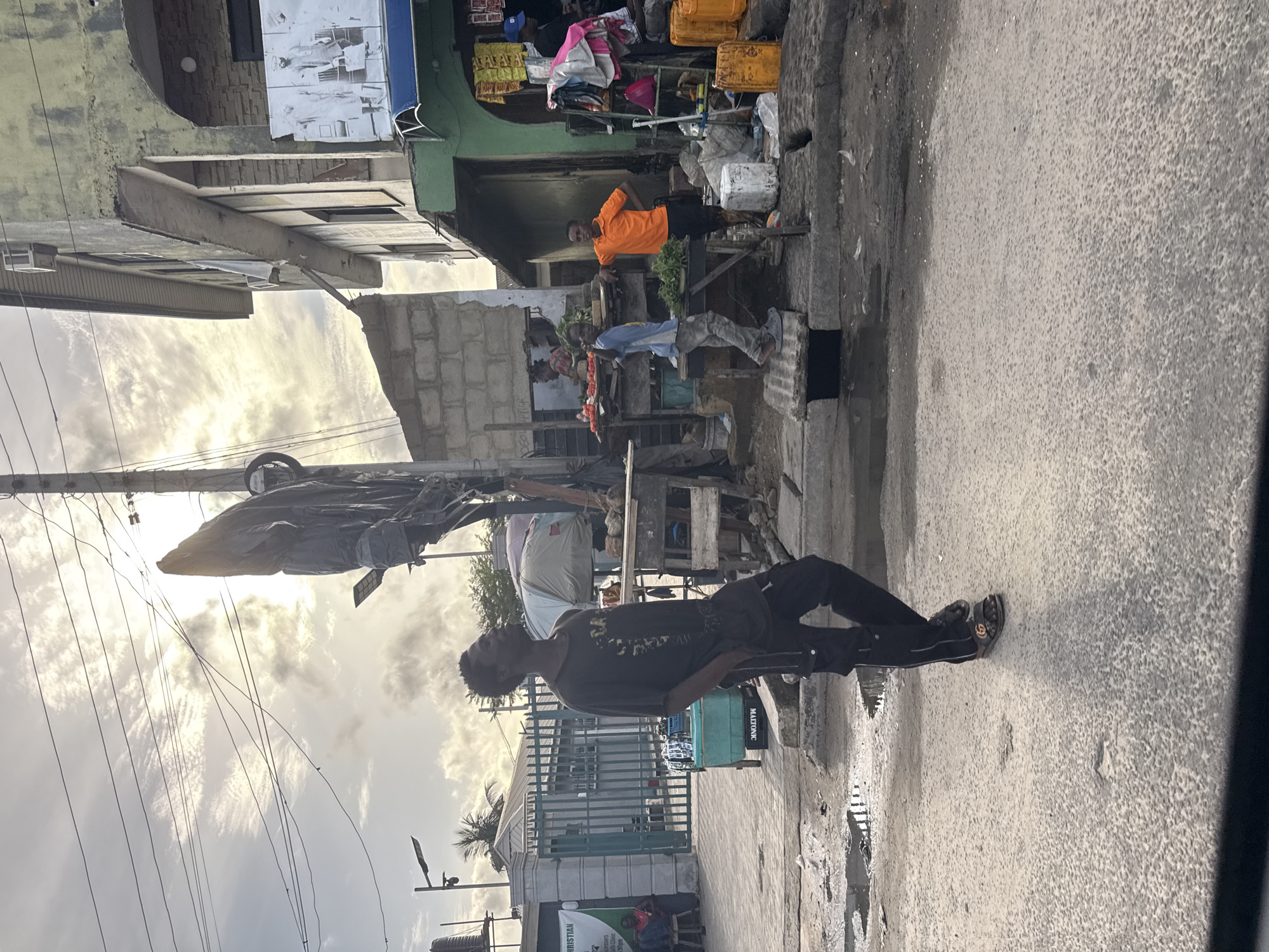 The image depicts a street scene in Ado, Nigeria. A young man wearing a dark t-shirt and pants is standing on the sidewalk. In the background, a market is visible with vendors selling produce and other goods. Other people stand and sit near the market. The buildings are a mix of concrete and block construction. Electrical wires crisscross overhead. The sky is overcast.