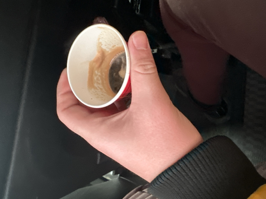 The image displays a light-skinned human hand holding a small, white paper cup with a red base, observed within the interior of a vehicle in the city of Ivanski, Bulgaria. The cup contains residual dark liquid, consistent with espresso or coffee, and light brown crema stains adhere to its inner walls. The subject's wrist is partially visible, encased by a dark, ribbed textile cuff. Below the hand and cup, a segment of dark brown fabric, likely part of trousers or a skirt, is discernible. The immediate background consists of dark, unidentifiable surfaces typical of an automobile's interior.