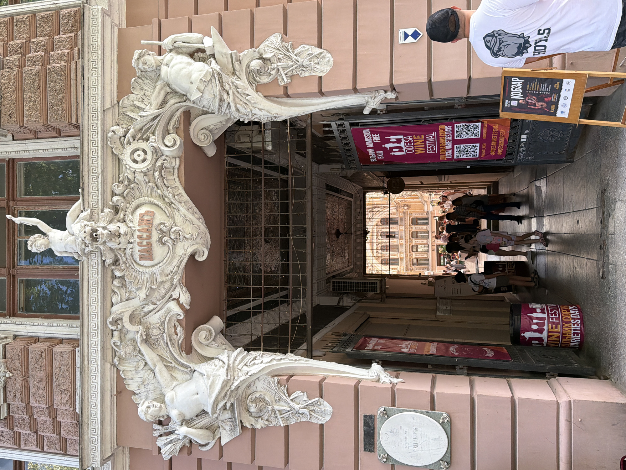 The image depicts the entrance to the Odesa Passage, a historic shopping arcade located in the city of Odesa, Ukraine. The facade of the building is adorned with ornate white stucco sculptures. Above the arched entrance, the word "ПАССАЖЪ" (Passage) is inscribed in a decorative cartouche. The entrance itself is an open archway leading to a covered walkway with a wrought-iron grille overhead.
In the foreground, to the right of the entrance, stands a man wearing a white t-shirt with a graphic of a bulldog and the word "HOYLS" printed on the back. He is looking away from the camera. To his left, an A-frame sign displays information about "КОБЗАР" with a picture of a musician.
Further inside the passage, several people are visible, including adults and children, walking or standing. Two large banners are hanging on the left side of the entrance, one of which advertises the "ODESA WINE FESTIVAL" and "LOCAL VARIETIES DAYS" with QR codes. Another banner is rolled up at the base of the entrance. A trash bin with similar wine festival imagery is also present. The architecture of the building is detailed and classical, with a pink-hued exterior. The overall scene suggests a vibrant public space, possibly during an event or festival.