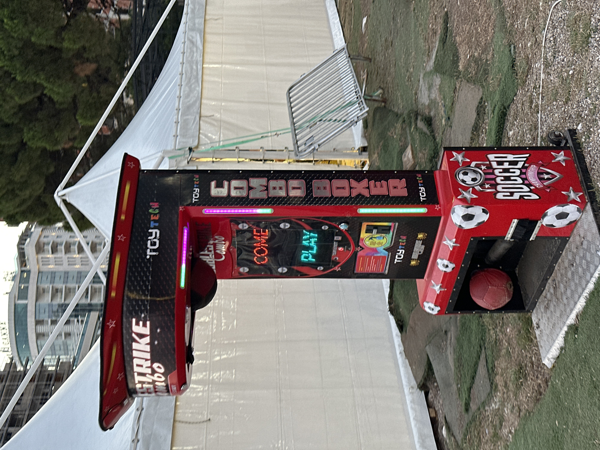 The image shows a "Combo Boxer Soccer" arcade game in an outdoor setting. The game is red and black with illuminated text and graphics. It features a punching bag area and a soccer ball slot. The game is set up on a grassy area with some gravel and artificial turf. In the background, there are white tents, a metal fence, trees, and a modern building. The scene is likely located in the city of Budva, Montenegro, possibly near a tourist attraction or event venue. There are no people visible in the image.