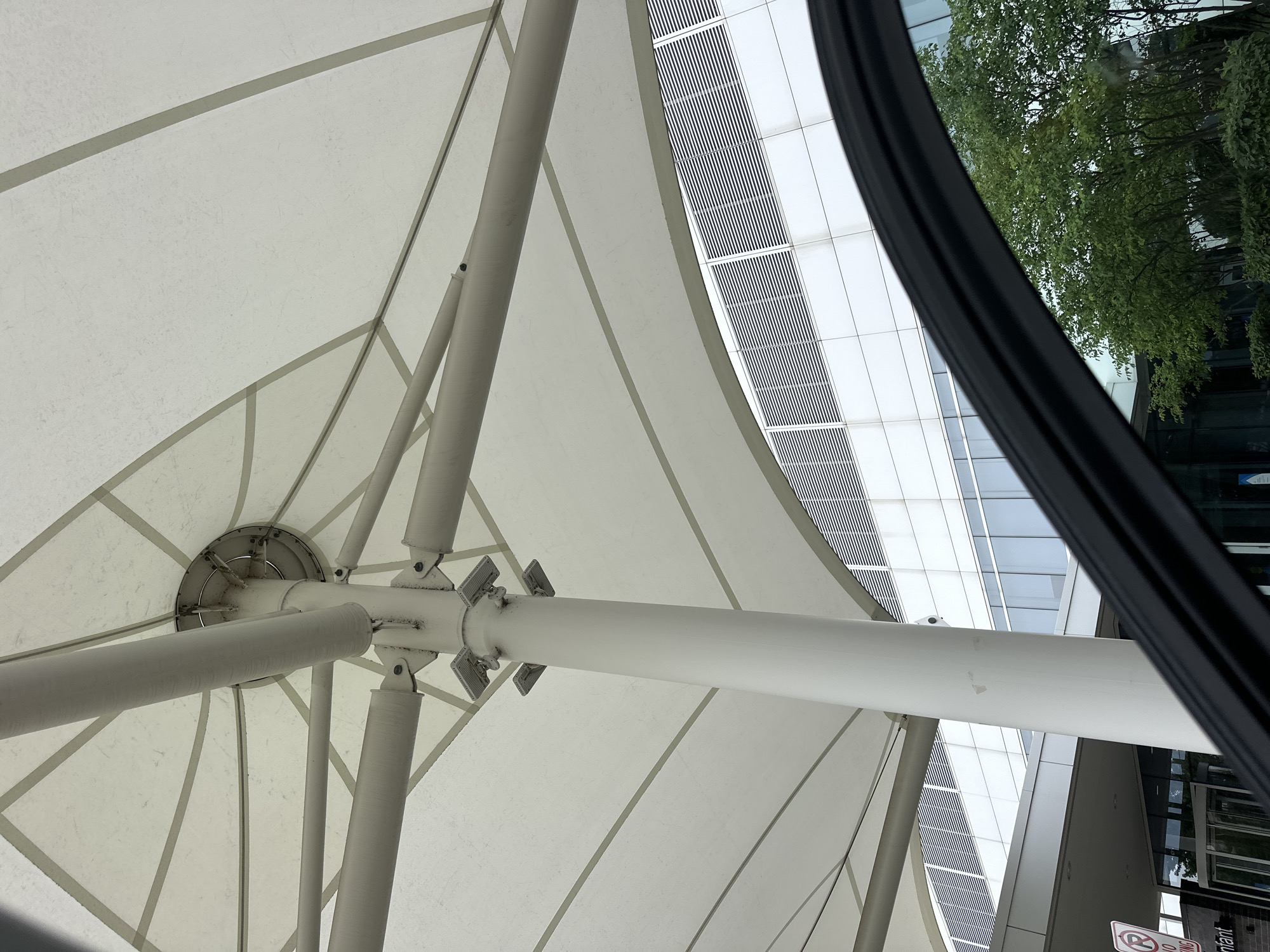 An upward angle shot shows the underside of a white tensile fabric structure supported by a central white pole and radiating white beams. The fabric is stretched taut and appears to be a canopy over an outdoor area. In the background, a modern building with a facade of glass windows and horizontal white louvers is visible. Trees with green foliage are reflected in the windows. A black car door frame or window trim curves into the foreground from the lower right. A red and white parking sign with a circle and diagonal line over a "P" is visible on the left side, along with the word "infant" on a sign below it, suggesting the location might be near a hospital or medical facility in Hoffman Estates, United States of America.