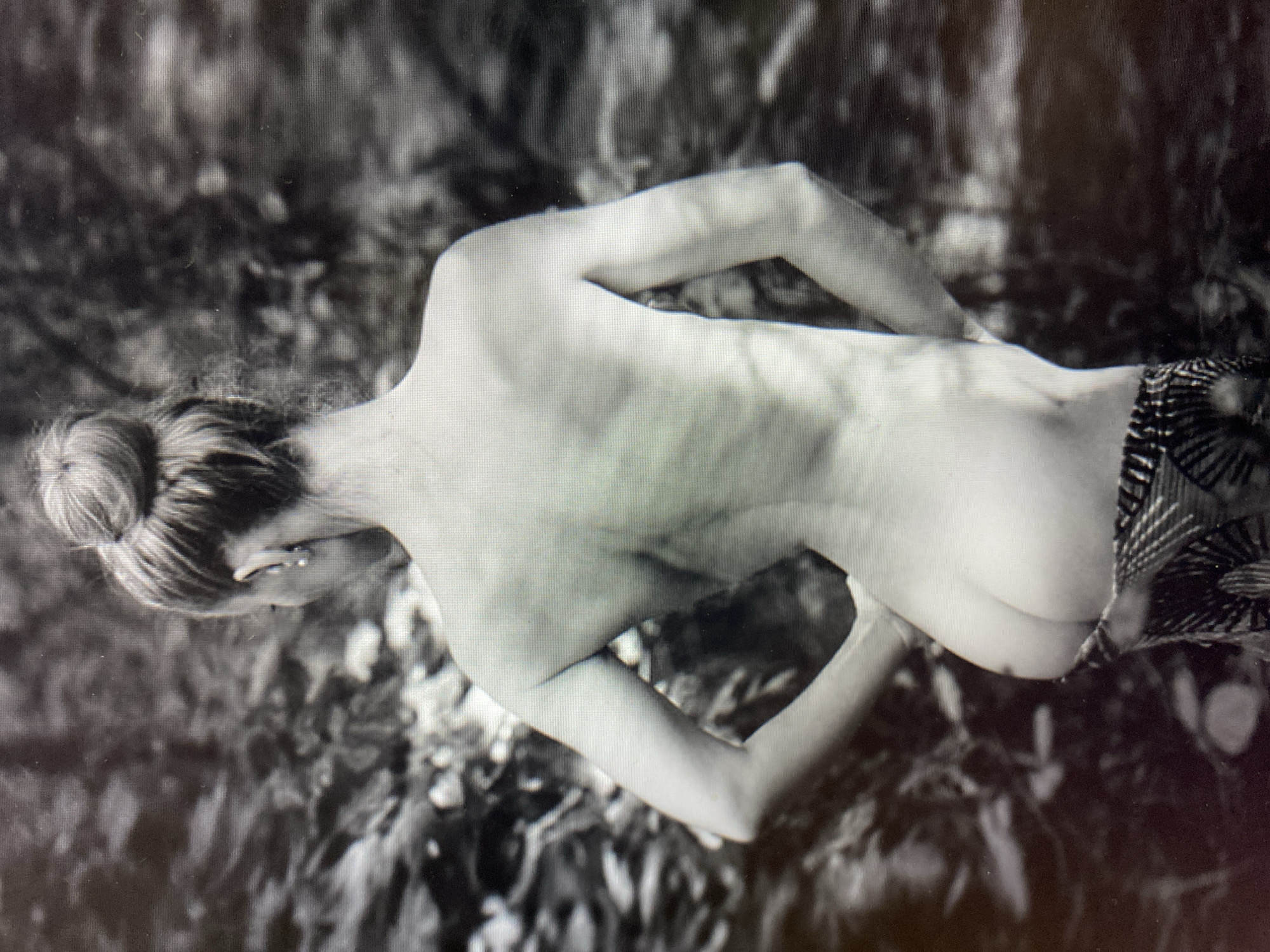This is a black and white, high-angle shot of a person from behind, seen from the waist up. The individual appears to be a woman with her hair pulled back into a bun. She is wearing patterned shorts or a skirt that covers her lower body. Her back is bare, revealing her spine and shoulder blades. Her arms are bent at the elbows, with her hands resting on her lower back. The background is out of focus and appears to be a natural setting with foliage. The lighting suggests it could be daytime, possibly outdoors, given the dappled light and shadow. The overall mood is serene and contemplative. There is no visible text in the image.