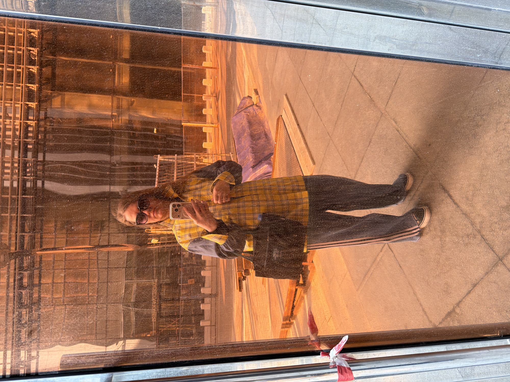 A woman wearing sunglasses, a yellow plaid shirt, black pants with stripes, and sneakers is taking a selfie. The image is a reflection in a window, showing a building under construction in the background. The location appears to be Moscow, Russia based on the Cyrillic lettering visible in the reflection. Other details include the woman holding a phone, a black tote bag, red and white striped tape near the bottom edge of the window, and tile flooring visible in the reflection of the ground.