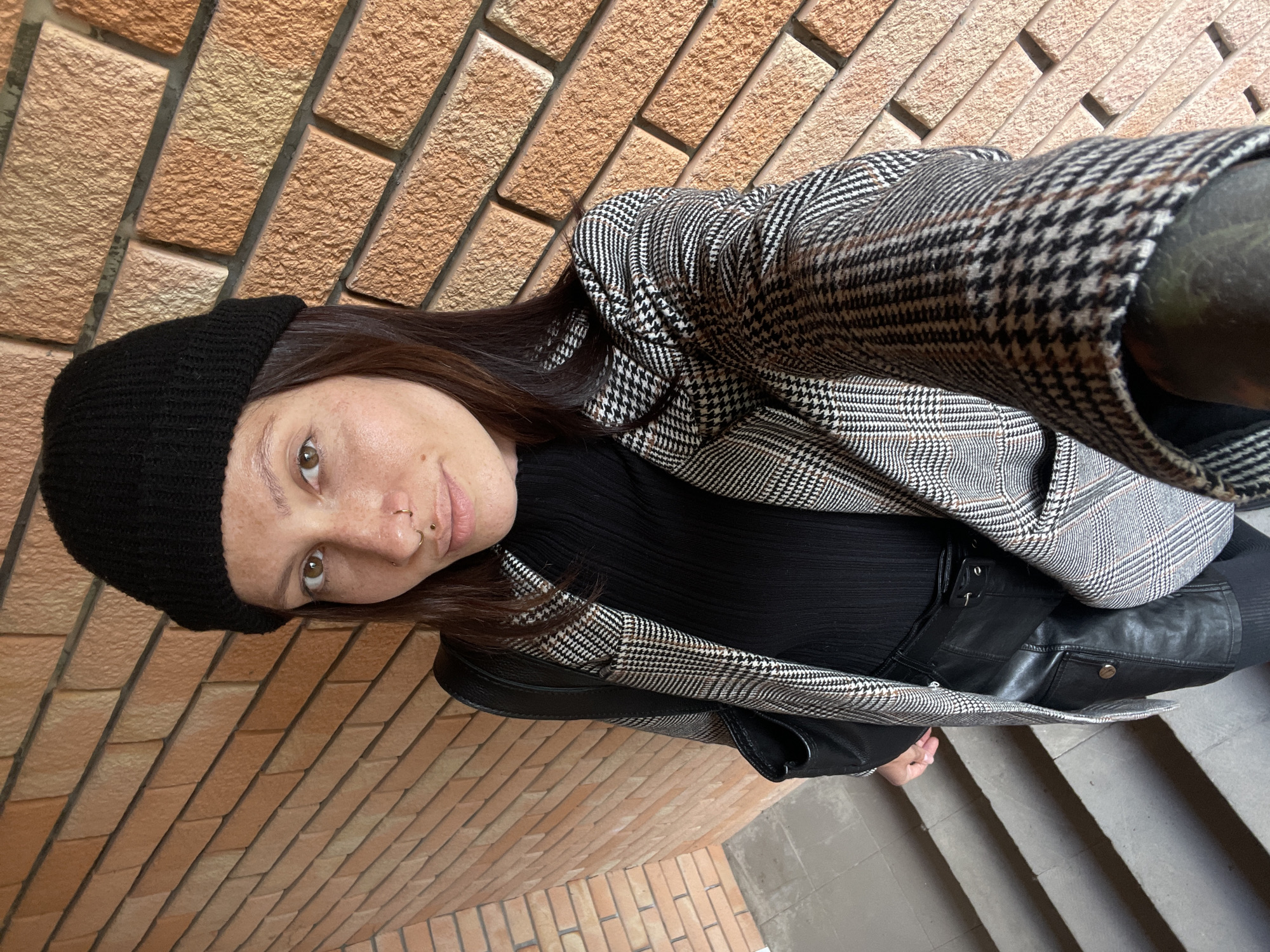 A young woman is posing in a photograph. She has brown hair, freckles and a nose ring. She is wearing a black beanie, a black top, a plaid jacket, and leather pants. She is standing on a set of grey concrete steps. Behind her is a light brown brick wall. The photograph was taken in Izhevsk, Russia.