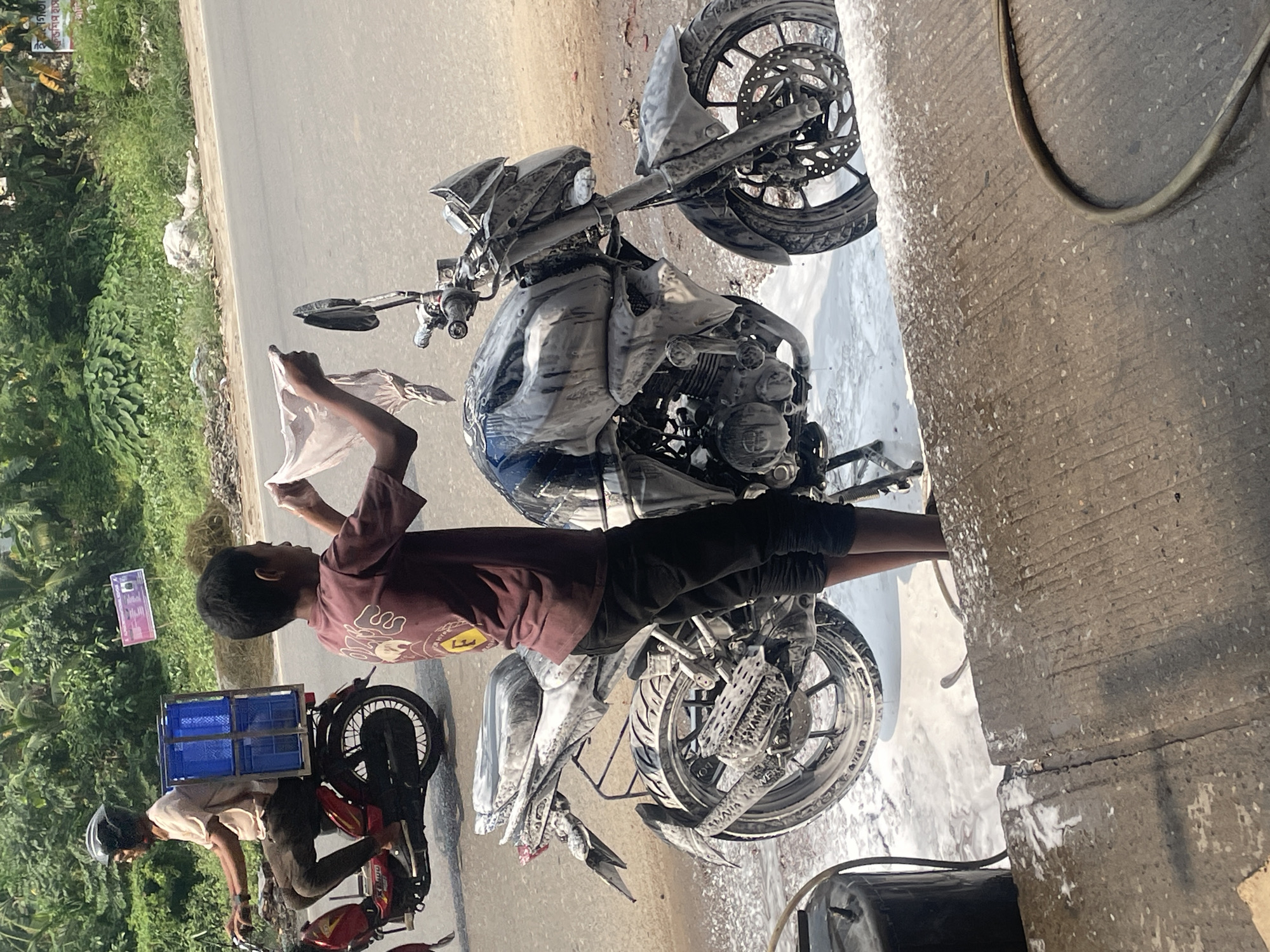 The image shows two motorcycles on a paved surface near vegetation, likely in Dhaka, Bangladesh. One motorcycle is covered in soap suds, being cleaned by a person holding a cloth. The second motorcycle, red and silver, is also parked nearby. A person on the second bike wears a helmet. A blue crate is attached to the rear of the second bike. The overall environment suggests an outdoor location, possibly a roadside area.