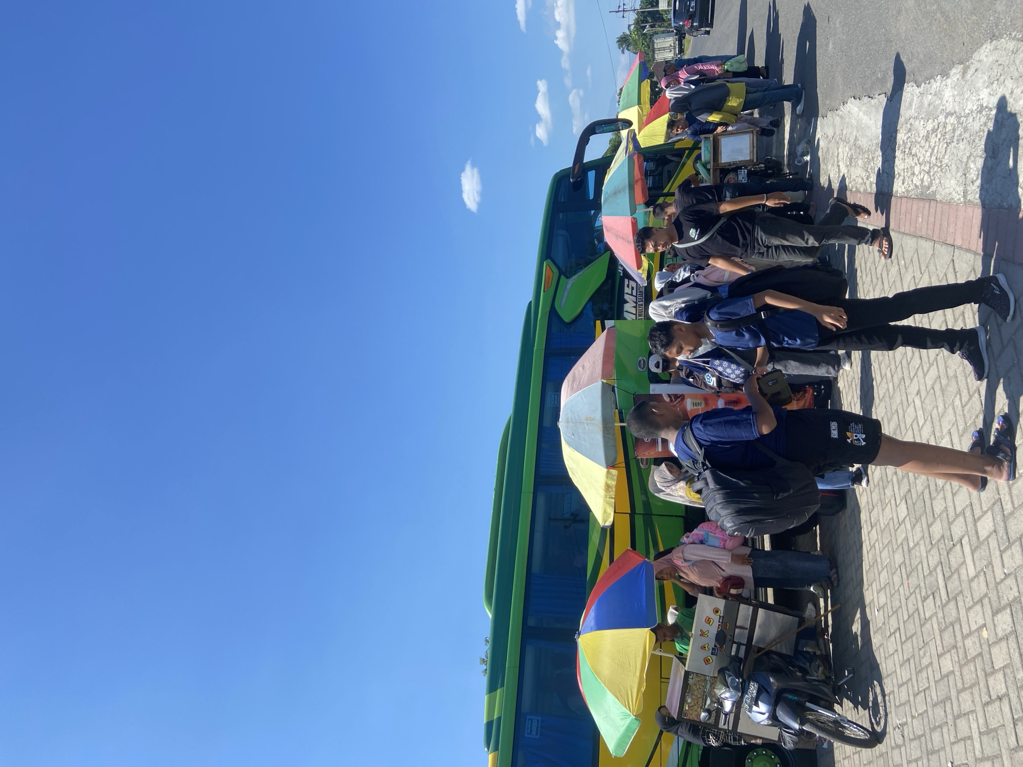 The image captures a street scene in Mangli, Indonesia. A green and yellow bus dominates the frame, parked along a paved road with a brick-lined edge. Several individuals are positioned adjacent to the bus, either standing or seated, under the shade of colorful umbrellas. The people appear to be diverse, with varying attire, including casual shirts, pants, and headscarves. Some carry bags, and others seem to be interacting with street vendors. A motorcycle with a food cart attached is visible, indicating a street food vending setup. The sky above is a clear, bright blue with scattered light clouds. The lighting suggests a sunny day, with shadows cast by the people and objects. In the background, a few vehicles are parked, and there are utility poles and trees visible, indicative of an urban or semi-urban environment.