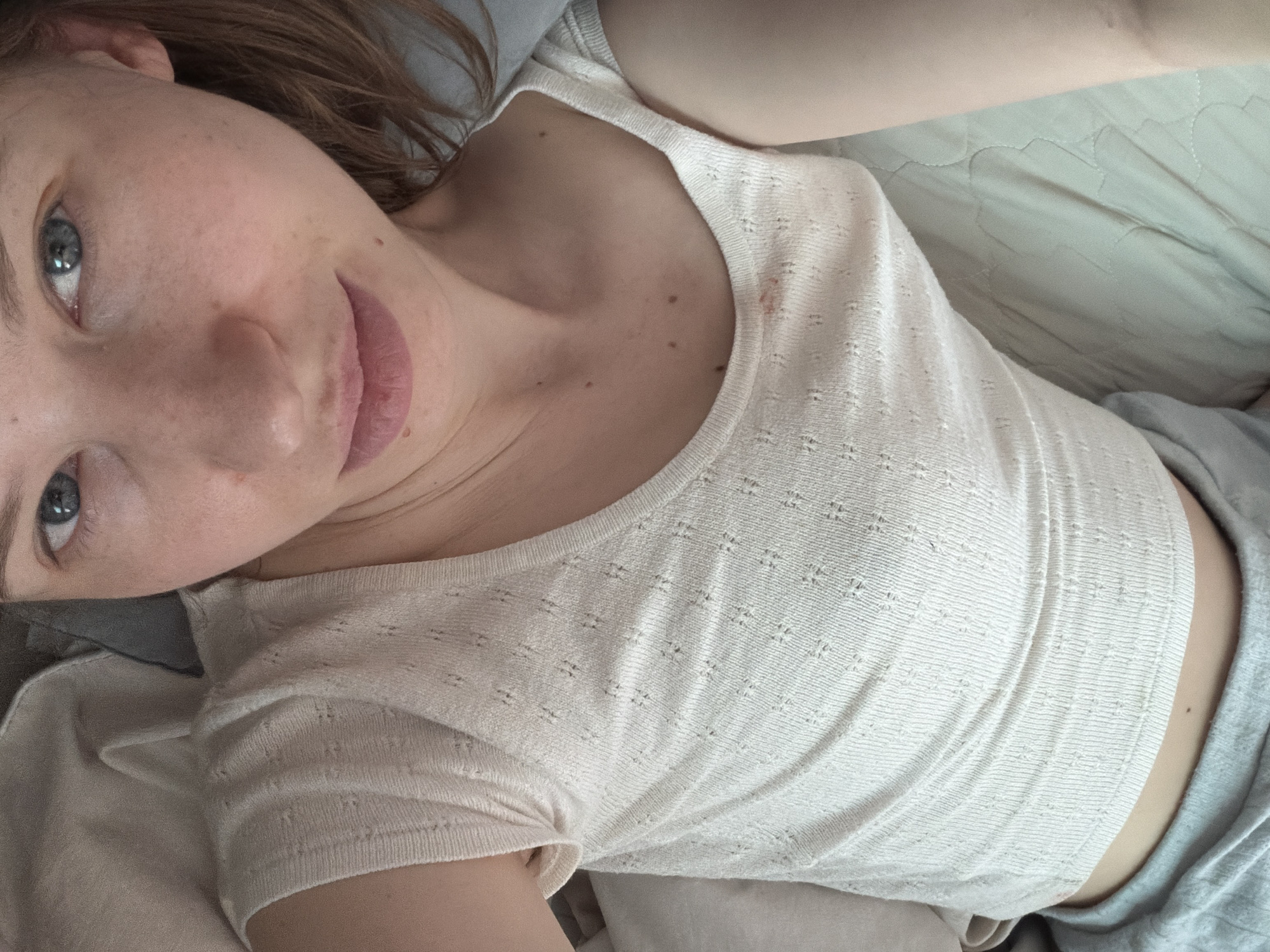 A woman with light skin, blue eyes, and light brown hair is lying in bed. She is wearing a light-colored, patterned tank top and grey shorts. The room is dimly lit, with natural light coming from the side. A light green quilted comforter is visible under her. The image appears to have been taken in a residential setting, possibly a bedroom. There are no other people or objects of significance in the frame. The location of Dongducheon-si, South Korea is not discernible from the image itself.