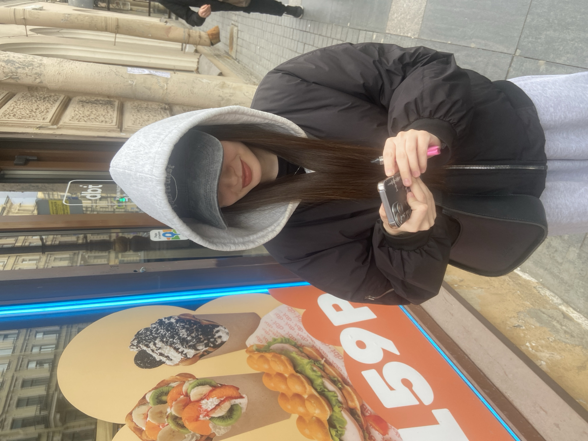 A young woman in a black puffy jacket and gray sweatpants, wearing a gray hooded cap, is taking a photo with her smartphone. She is standing in front of a shop window showcasing images of bubble waffles with various toppings. The pricing of these waffles is displayed prominently as "159P" (presumably Russian Rubles). The shop window has a bright blue neon light strip along the top edge. Reflections of buildings are visible in the glass. The location appears to be in Saint Petersburg, Russia, based on the architecture and the currency displayed. In the background, part of another person can be seen walking down the street.