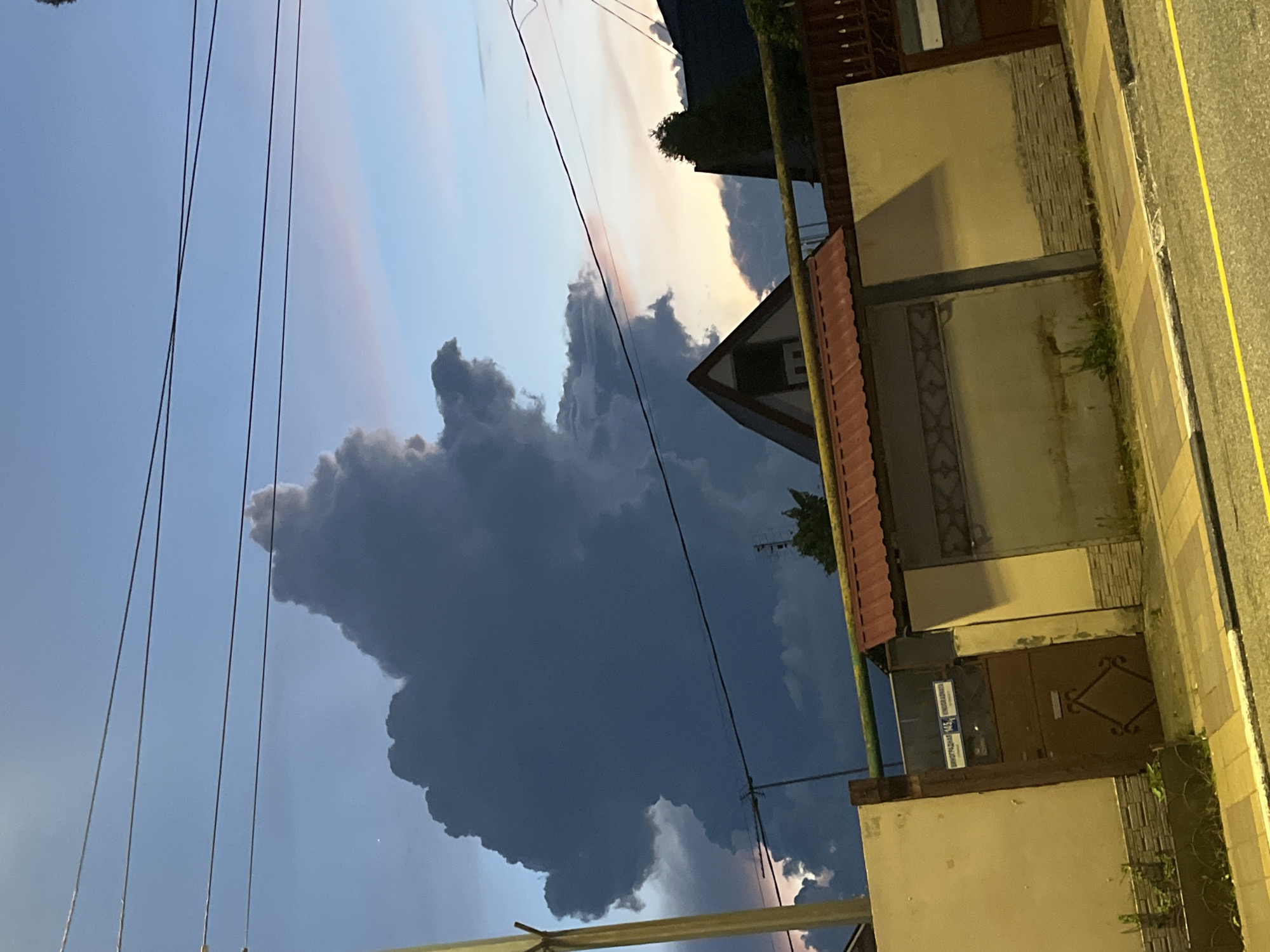 This image depicts a cloudy sky over a residential area in Sochi, Russia, possibly during dusk or dawn. A large, dark cloud formation dominates the upper left portion of the frame, casting a dramatic shadow over the scene. In the foreground, a fence with a metal gate and two signs with Cyrillic text is visible. A house with a red-tiled roof and an attic window is situated behind the fence. Power lines stretch across the sky, and a street with yellow markings runs along the bottom of the image. The overall atmosphere is subdued, with the dim lighting suggesting either early morning or late evening. There are no people or animals visible in the image.