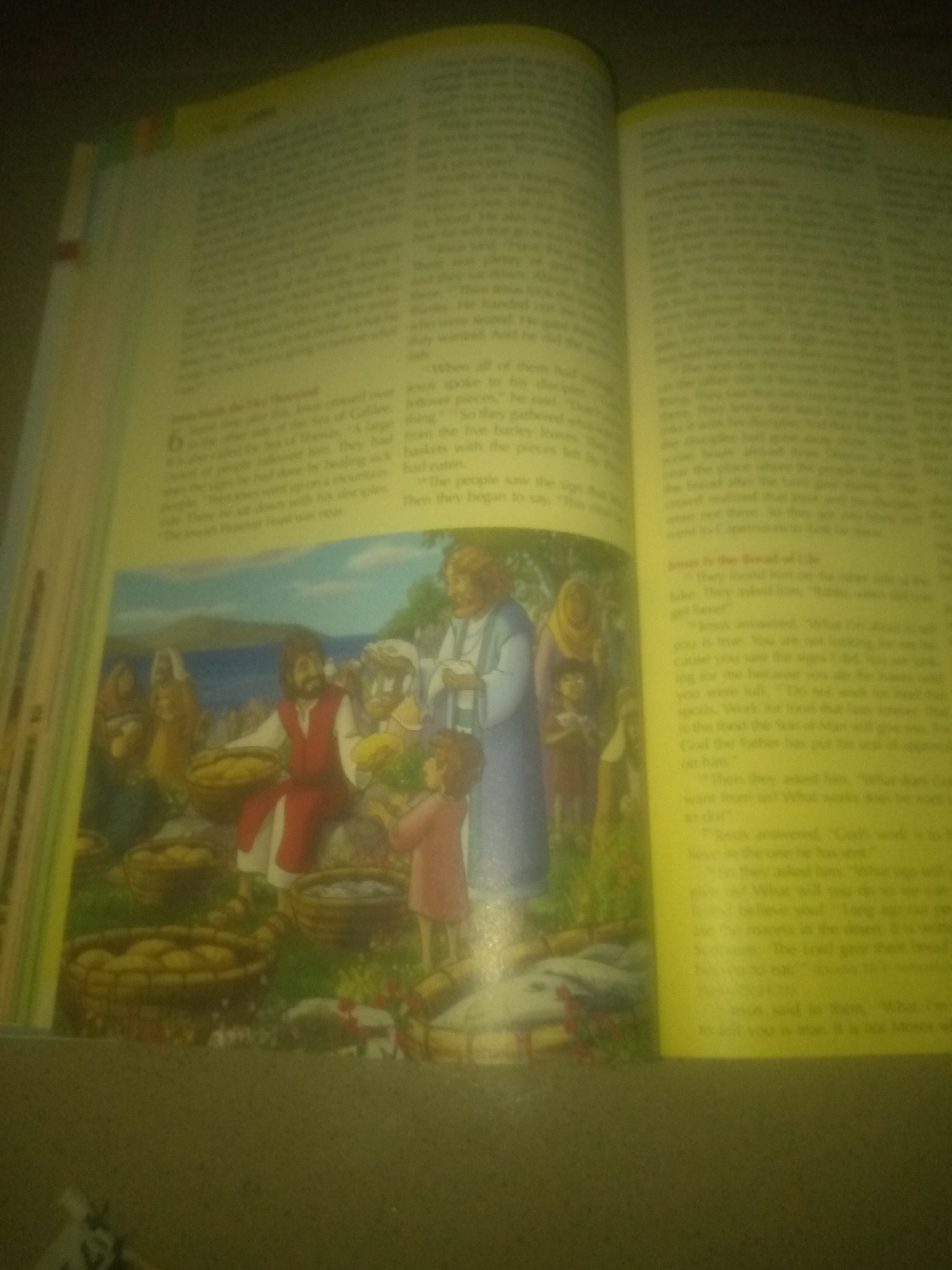 The image shows an open book, likely a Bible, with text and an illustration. The illustration depicts a biblical scene with Jesus and a crowd of people, likely the feeding of the multitude. Jesus is shown in the center, distributing bread from baskets. There are many people gathered around him, and baskets of bread and fish are visible in the foreground. The text on the left page appears to be from the Gospel of John, discussing the feeding of the five thousand. The text on the right page discusses Jesus being the Bread of Life. The lighting suggests the image was taken indoors with artificial light. There is no information about Jauro Yinu, Nigeria in the image.