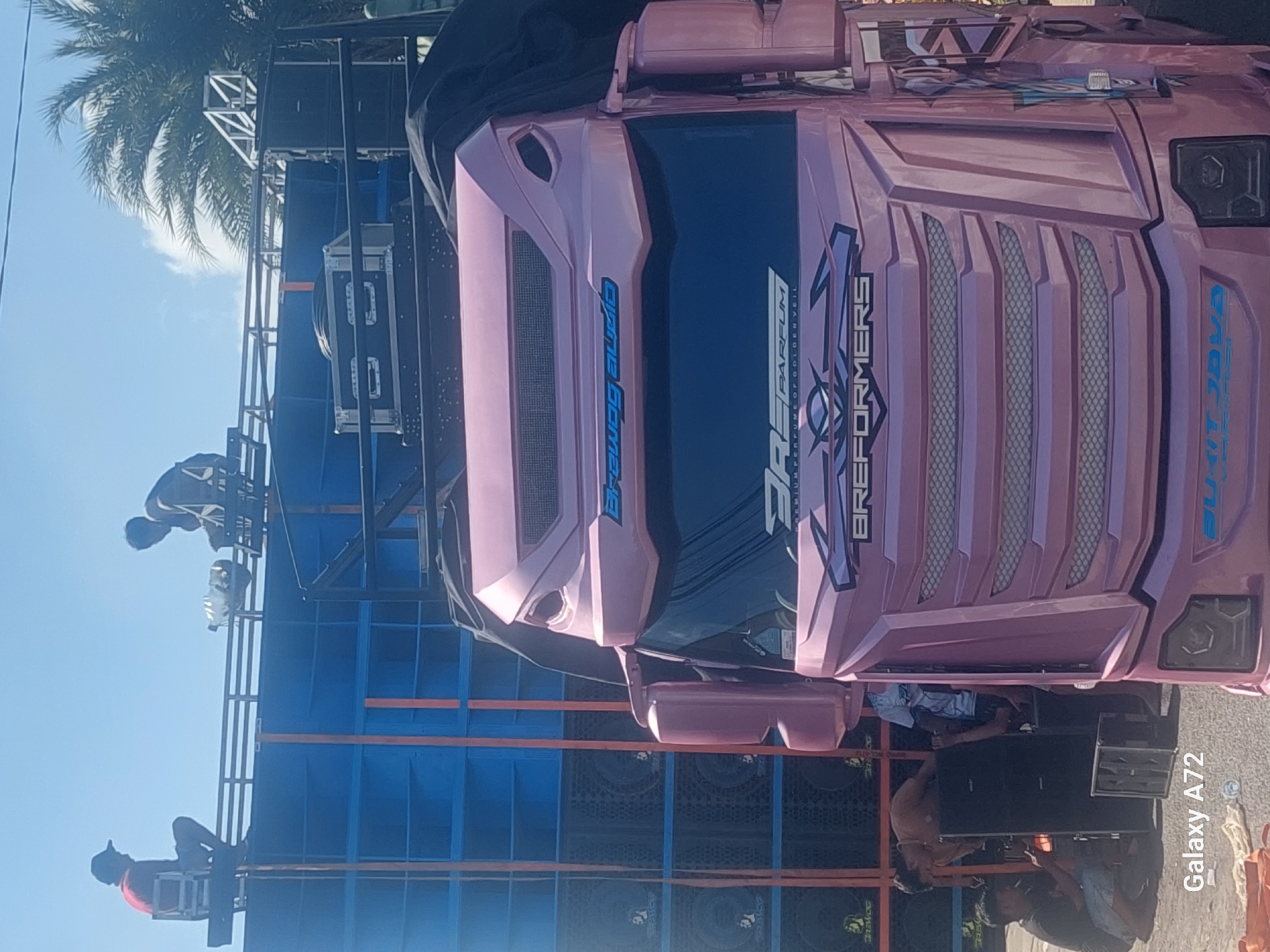 In Sengguruh, Indonesia, a pink truck with custom graphics, including "BRAVOZ AUDIO" and "BREFORMERS" on the front, is parked in front of a large, blue speaker system. Several individuals are present. Two men are seated on a metal structure atop the speaker system, with one wearing a red shirt and a cap, and the other in a dark shirt. Below the speaker system, several people are actively working. One man in a black t-shirt and shorts is crouching and appears to be connecting wires to a black speaker. Another person, partially visible, is also near the speakers. In the background, palm trees are visible against a clear blue sky. The overall scene suggests an event or setup involving sound equipment.